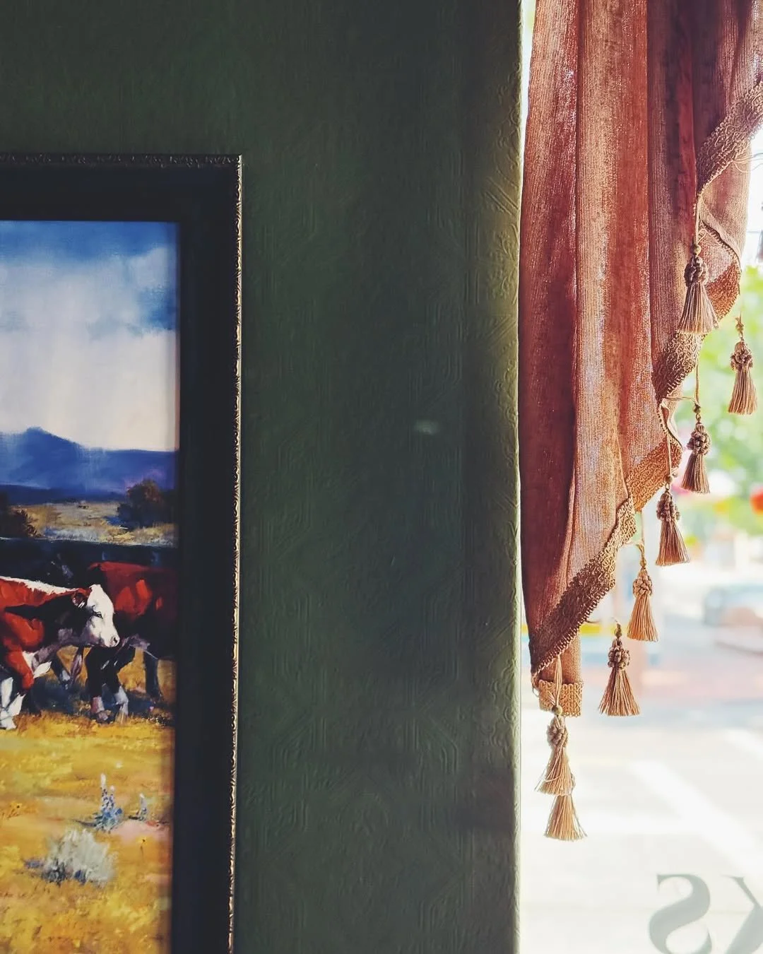 Part of a framed landscape painting on a green wall, with a pink curtain featuring tassels hanging next to a window with sunlight coming through.
