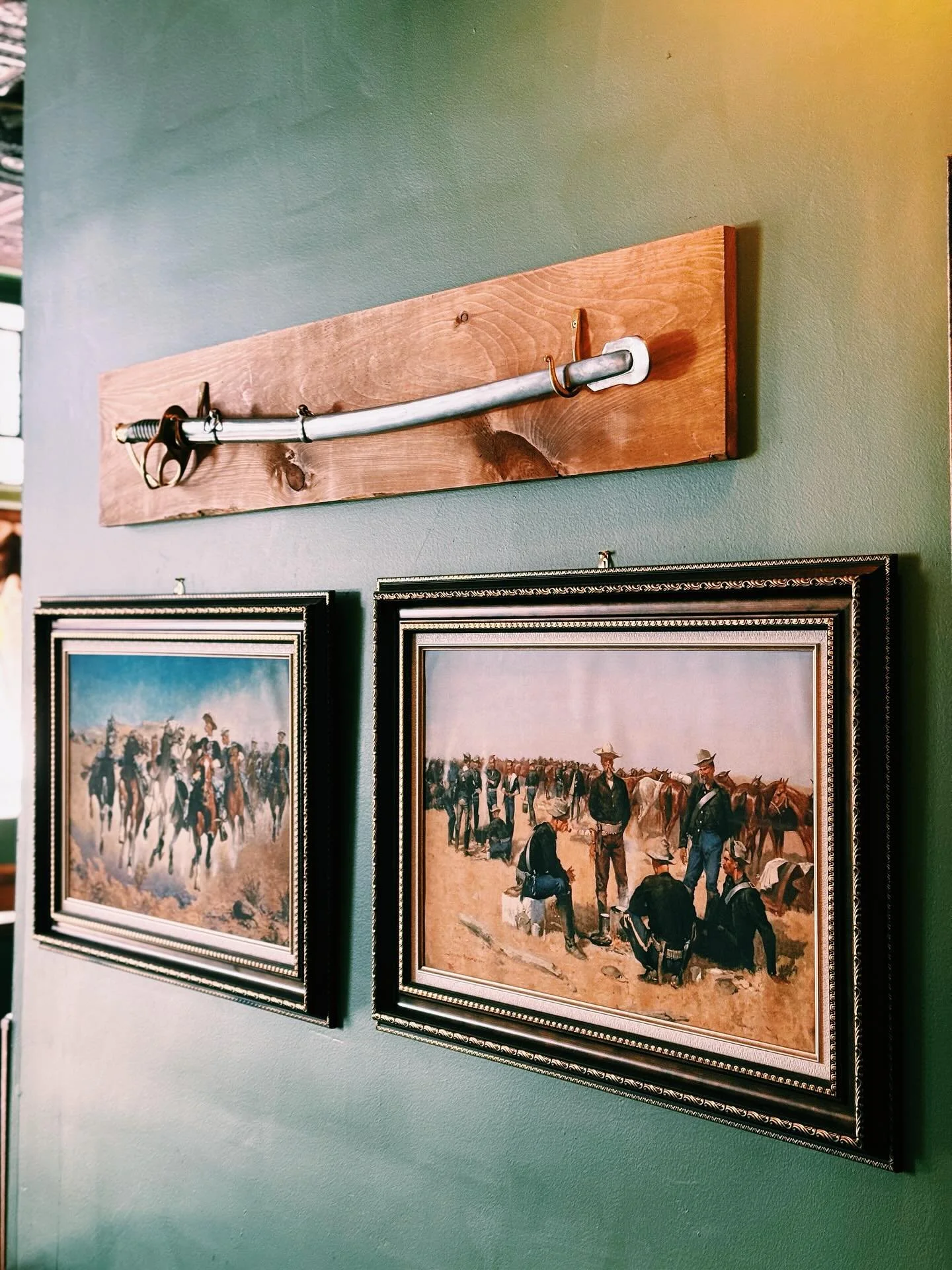 A decorative display on a green wall featuring a sword mounted on a wooden plaque above two framed paintings of historic scenes with people and horses.