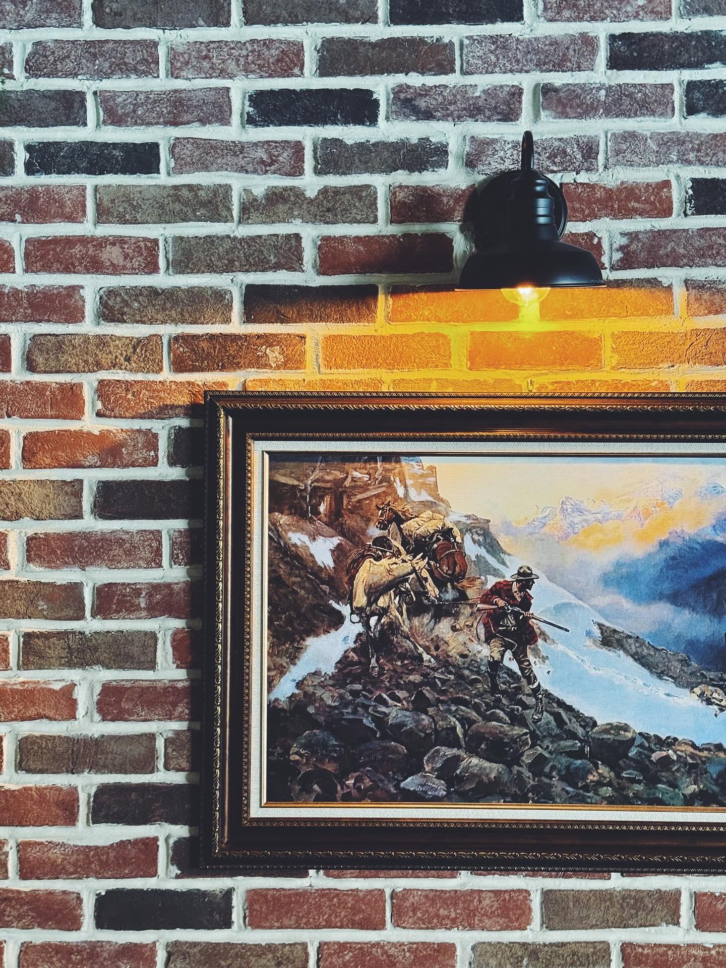 Historical painting of soldiers crossing a rocky mountain path with snow, mountains in the background, under a partly cloudy sky, framed on a brick wall."