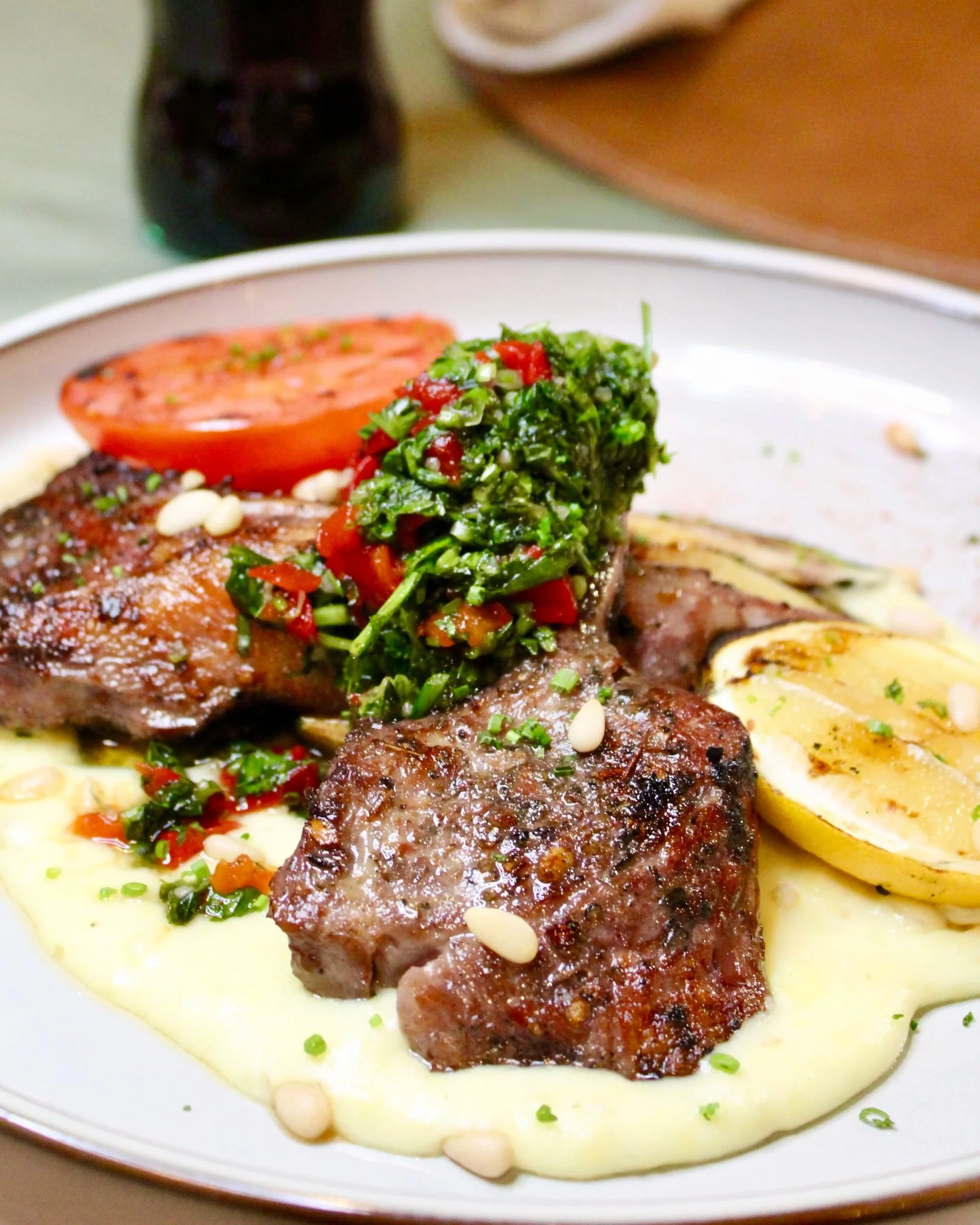 A plate of grilled meat served with mashed potatoes, topped with a green herb mixture and pine nuts, accompanied by a grilled lemon half and a slice of tomato.