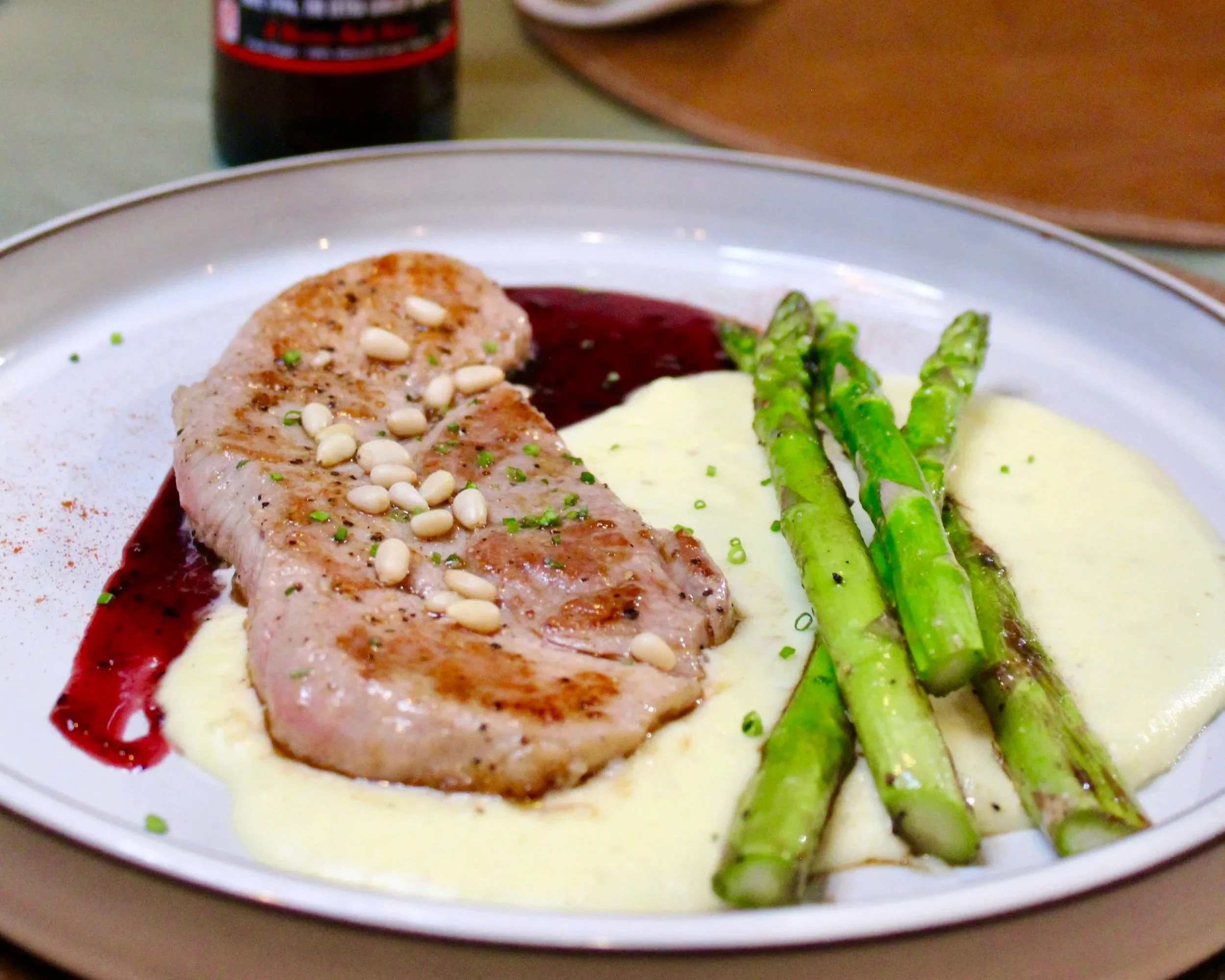 A plate with a grilled meat steak topped with pine nuts, served with green asparagus spears and creamy mashed potatoes, garnished with chopped chives and a side of red sauce.