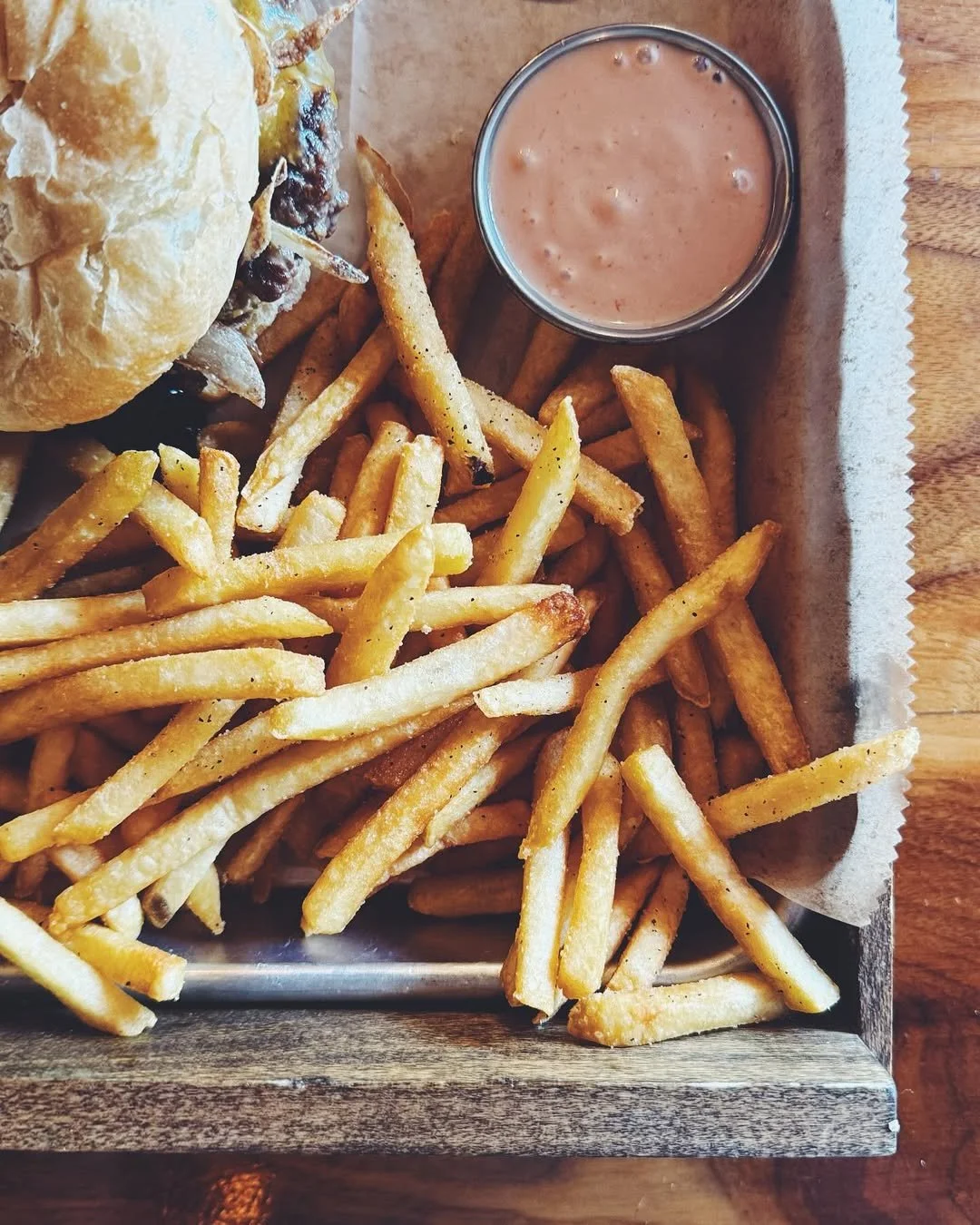 A metal tray filled with French fries, a burger partially visible, and a small cup of dipping sauce on a wooden table.