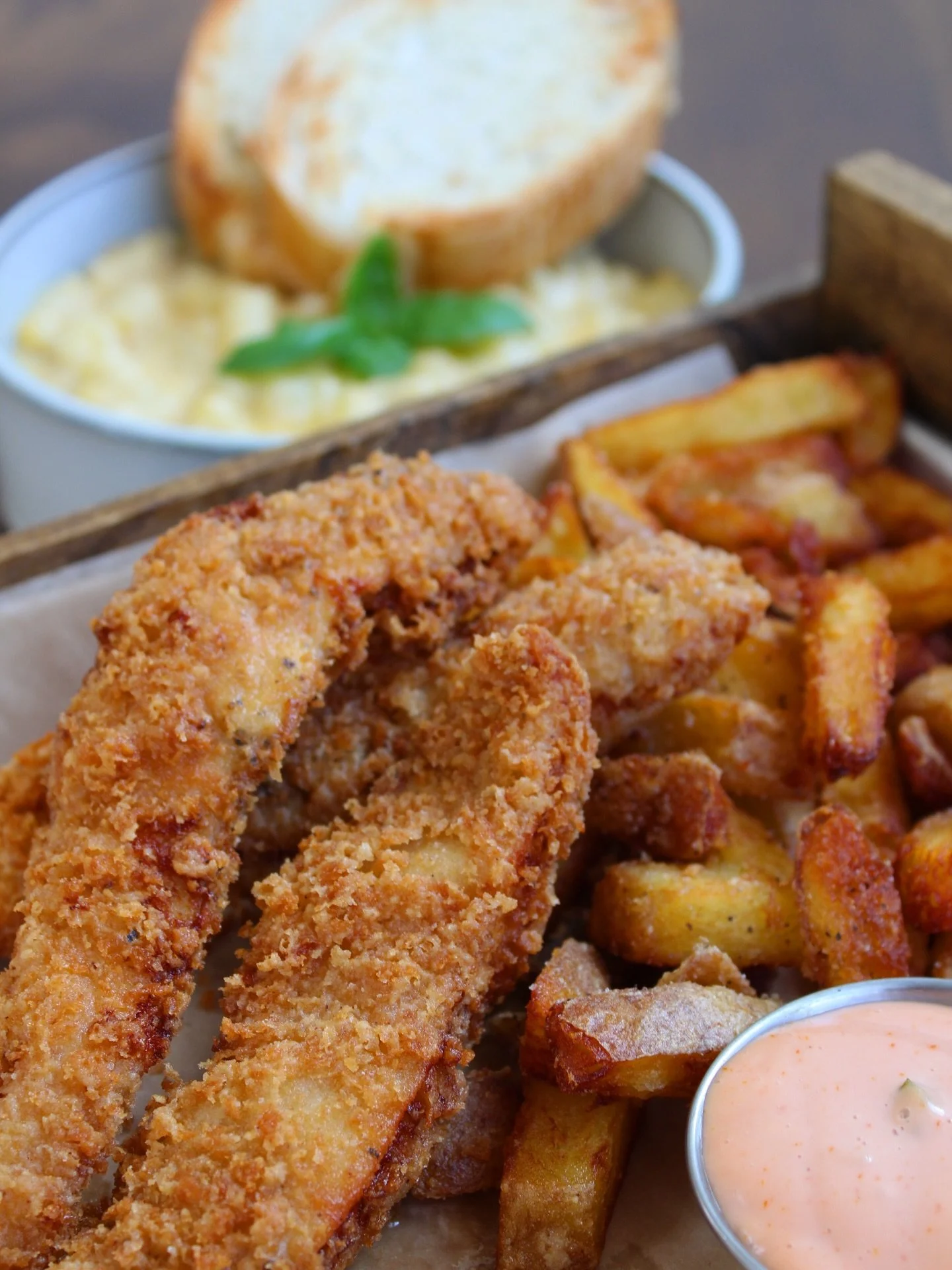 Close-up of fried fish fillets, crispy potato wedges, a side of pink dipping sauce, and a small bowl of macaroni and cheese with a garlic bread slice on top, served on a wooden tray.