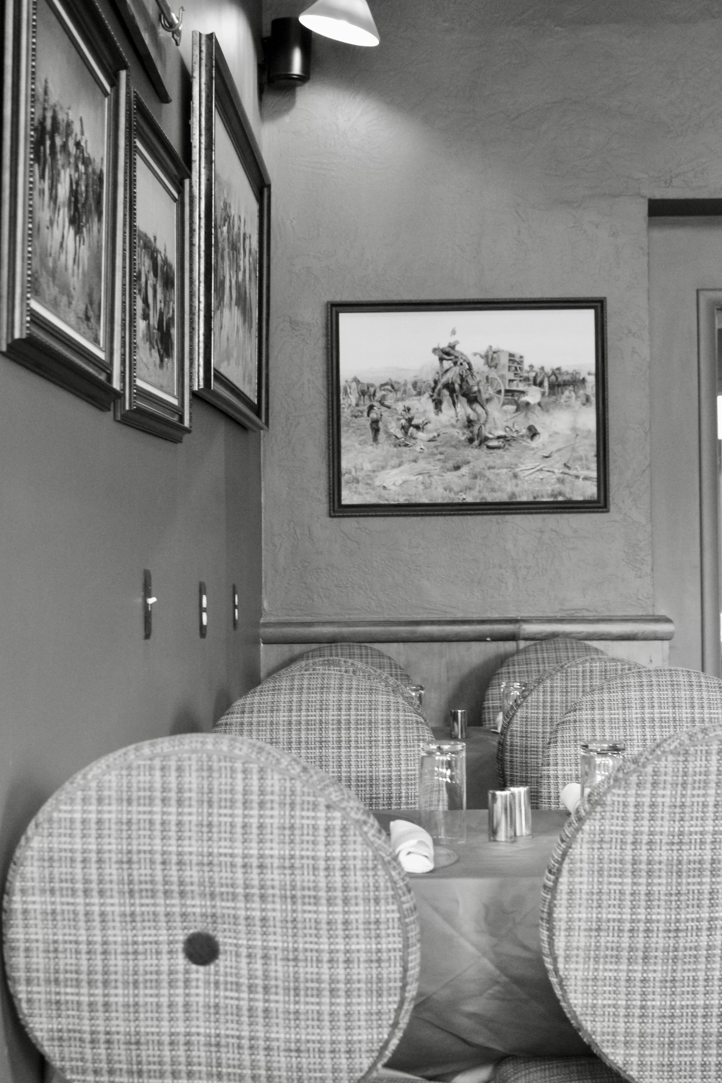 A black and white photo of a restaurant interior with a round table set with glasses and napkins, upholstered chairs, decorated wall with framed paintings, and a textured wall with a framed picture of a historic scene.