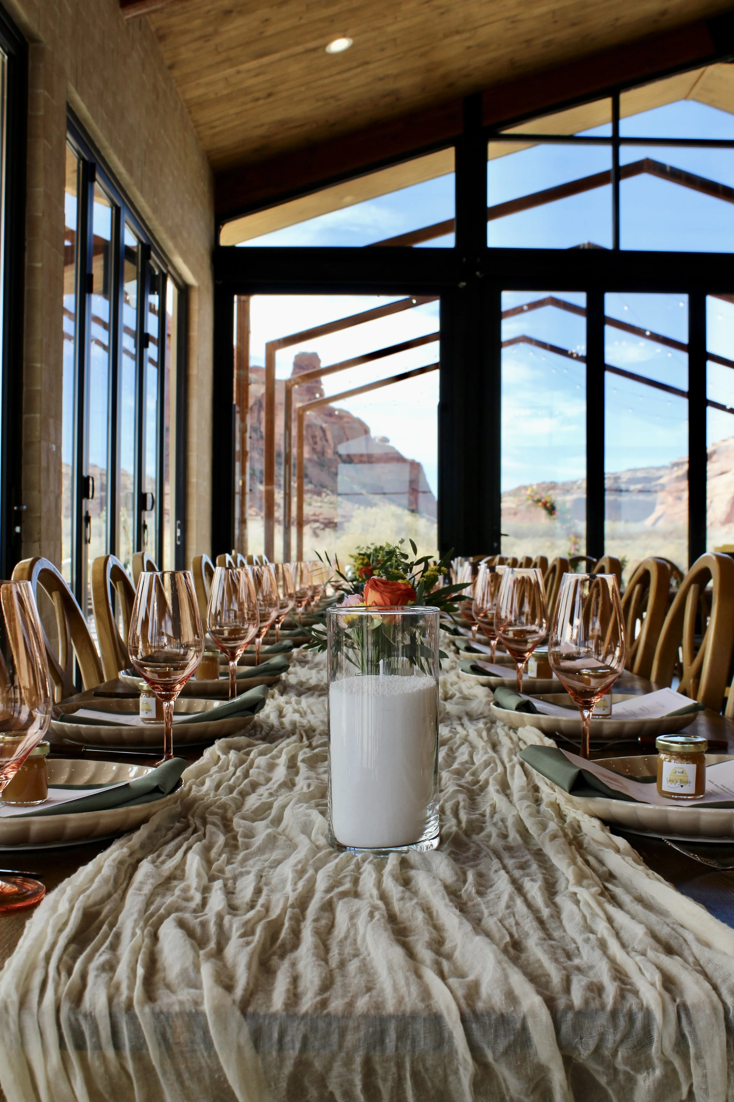 Set dining table with wine glasses, napkins, and a floral centerpiece inside a windowed room overlooking desert rock formations.