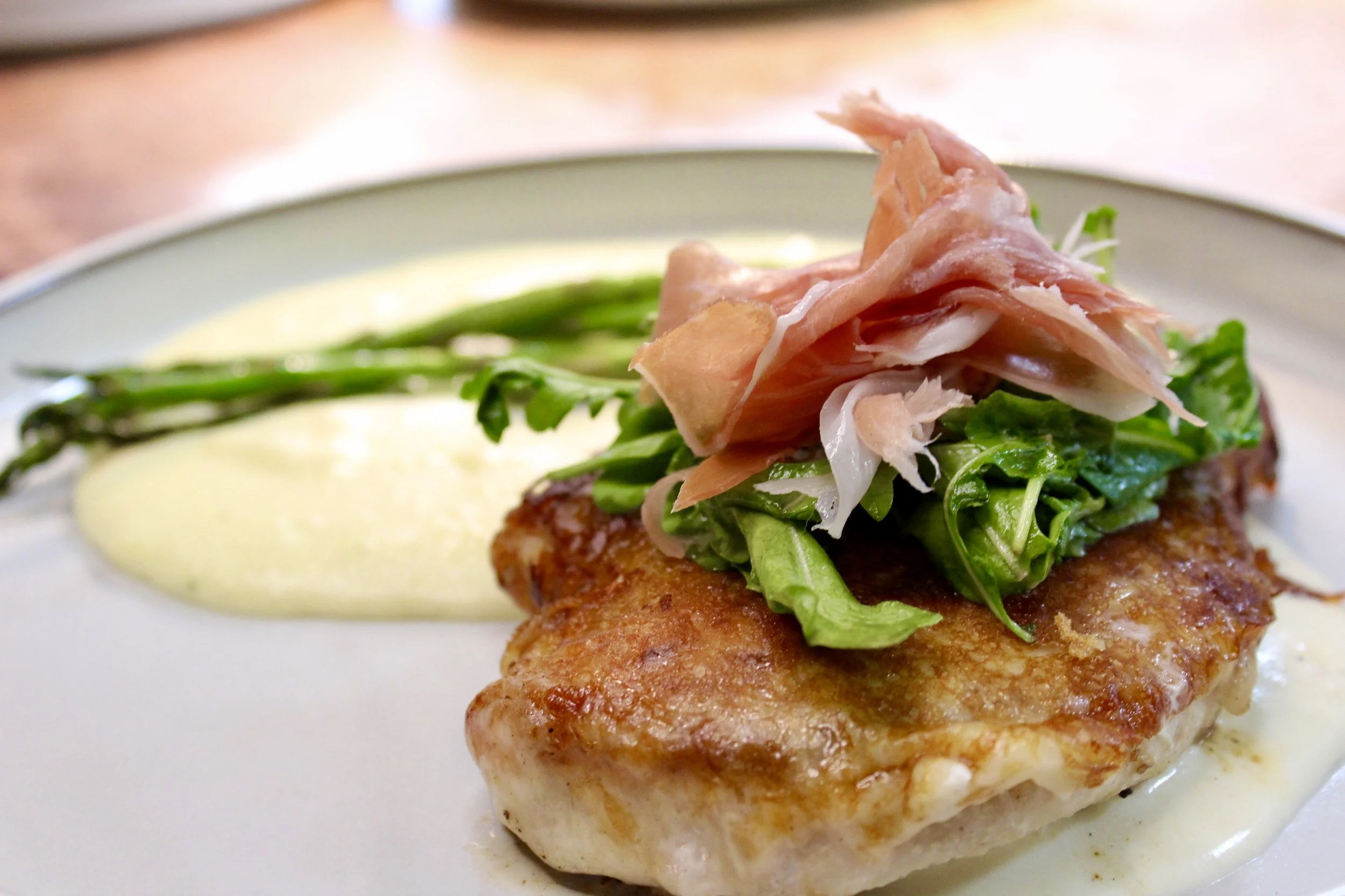 Close-up of a plated dish featuring a piece of cooked fish topped with green leafy vegetables, slices of prosciutto, and a side of asparagus, with a creamy yellow sauce underneath.