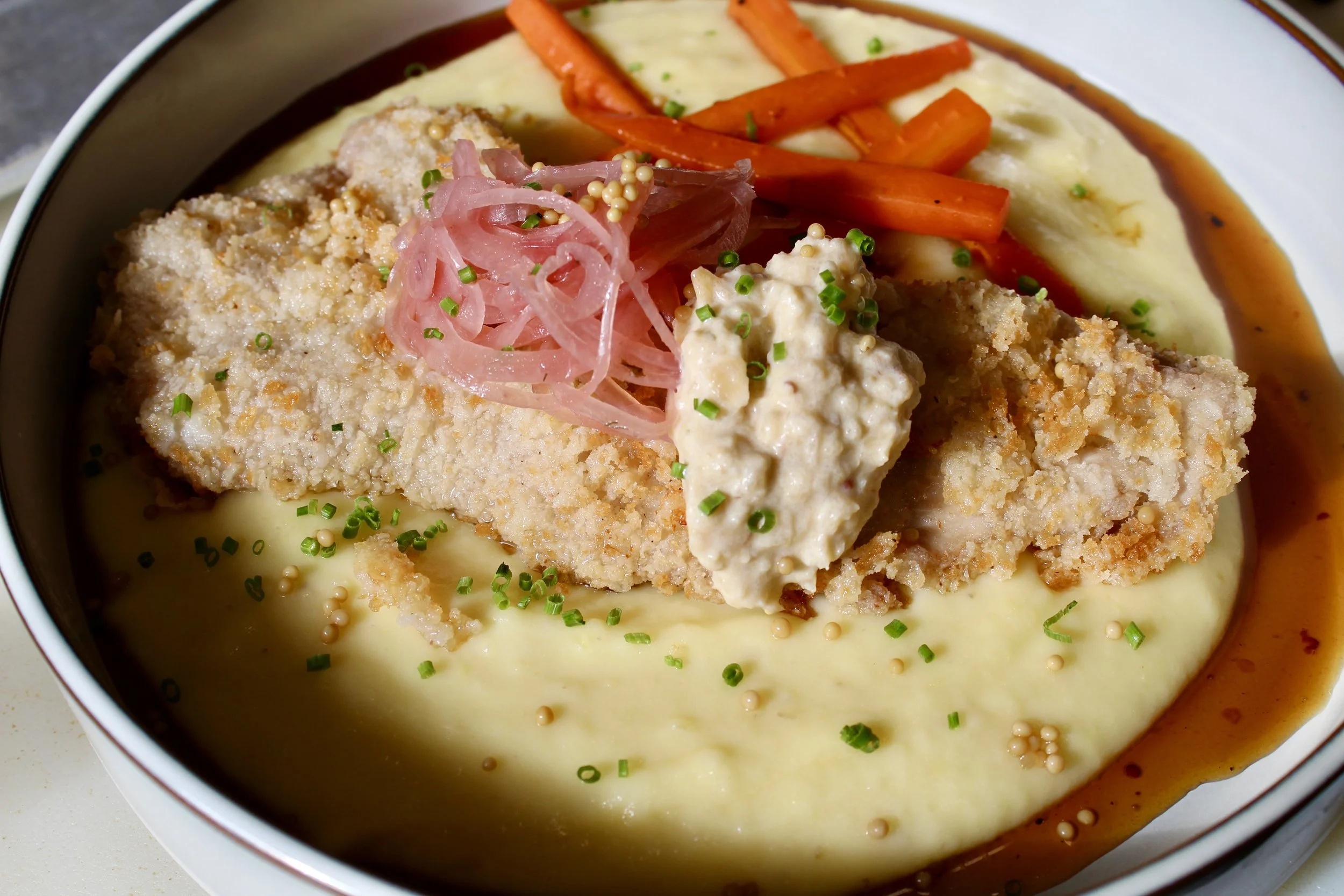 Breaded fish fillet served over mashed potatoes with gravy, topped with pickled onions, a dollop of tartar sauce, and garnished with chopped chives, alongside cooked carrots.