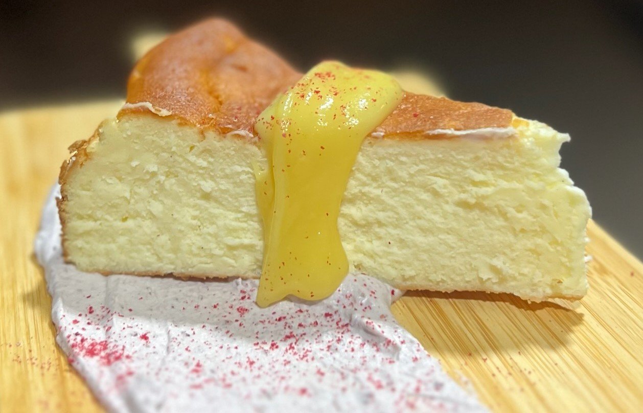 Slice of cheesecake with lemon curd topping on a wooden board, garnished with pink powder and red spice.