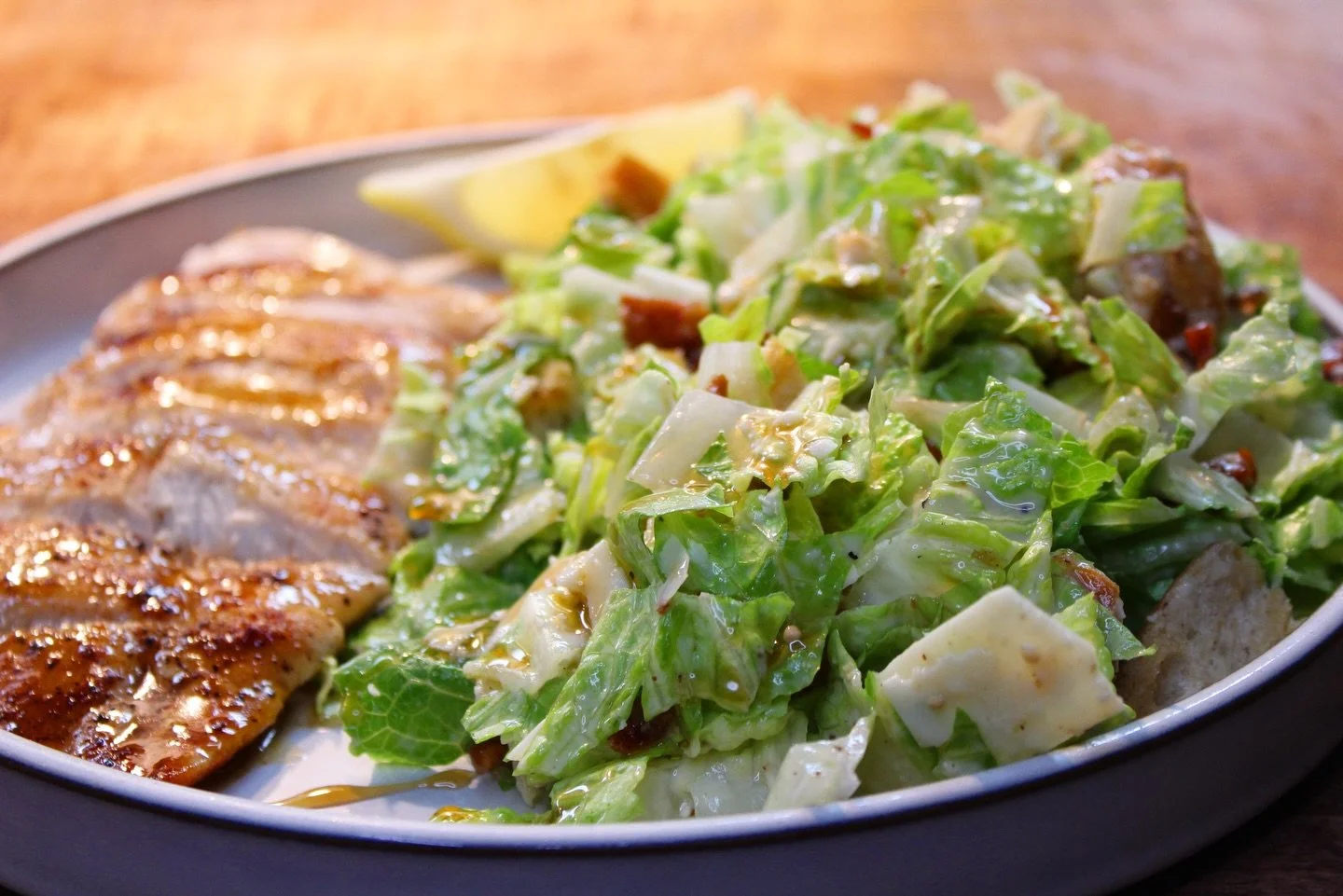 Grilled chicken served with a side Caesar salad topped with dressing and lemon wedge on a gray plate.