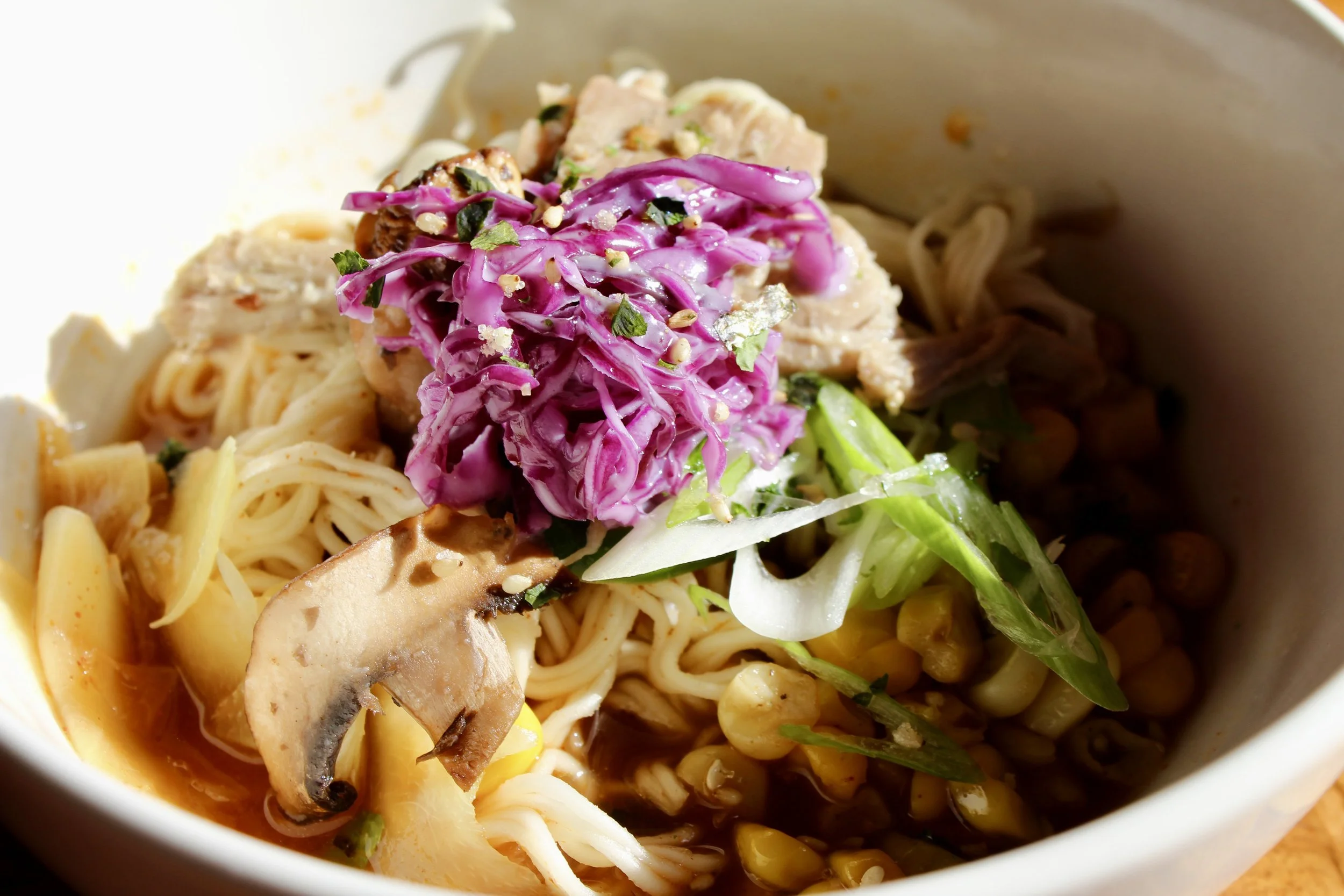 Close-up of a bowl of ramen noodles topped with purple cabbage slaw, sliced chicken, mushrooms, corn, and chopped green onions.