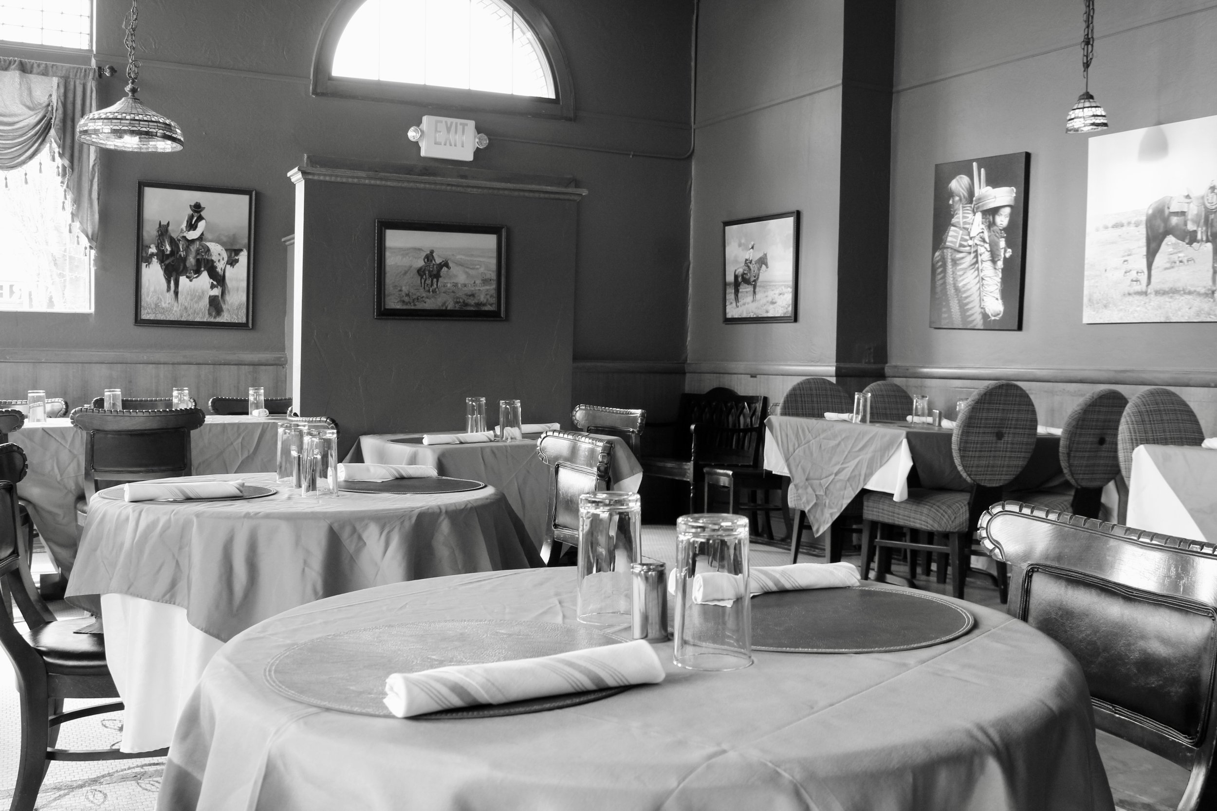 Interior of a restaurant with round and rectangular tables, set with glasses, napkins, and tablecloths. The walls are decorated with framed pictures of horses and people with ranching gear. There are windows and hanging light fixtures.
