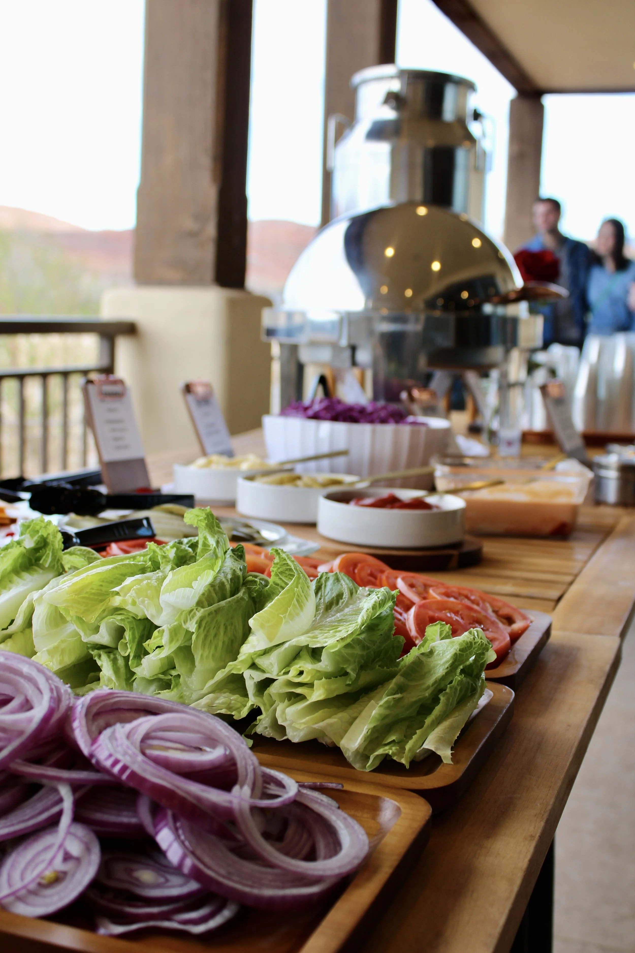 Buffet table with fresh vegetables including sliced onions, lettuce, tomatoes, and other salad ingredients at an outdoor event.