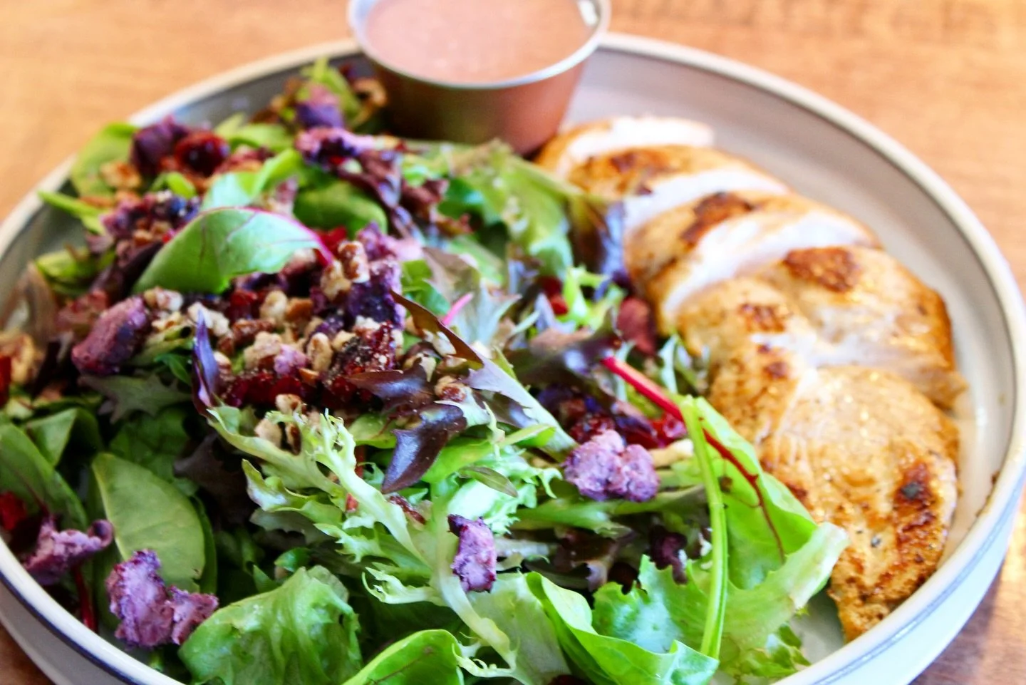 Mixed green salad with sliced grilled chicken breast and a side of dressing in a small bowl.