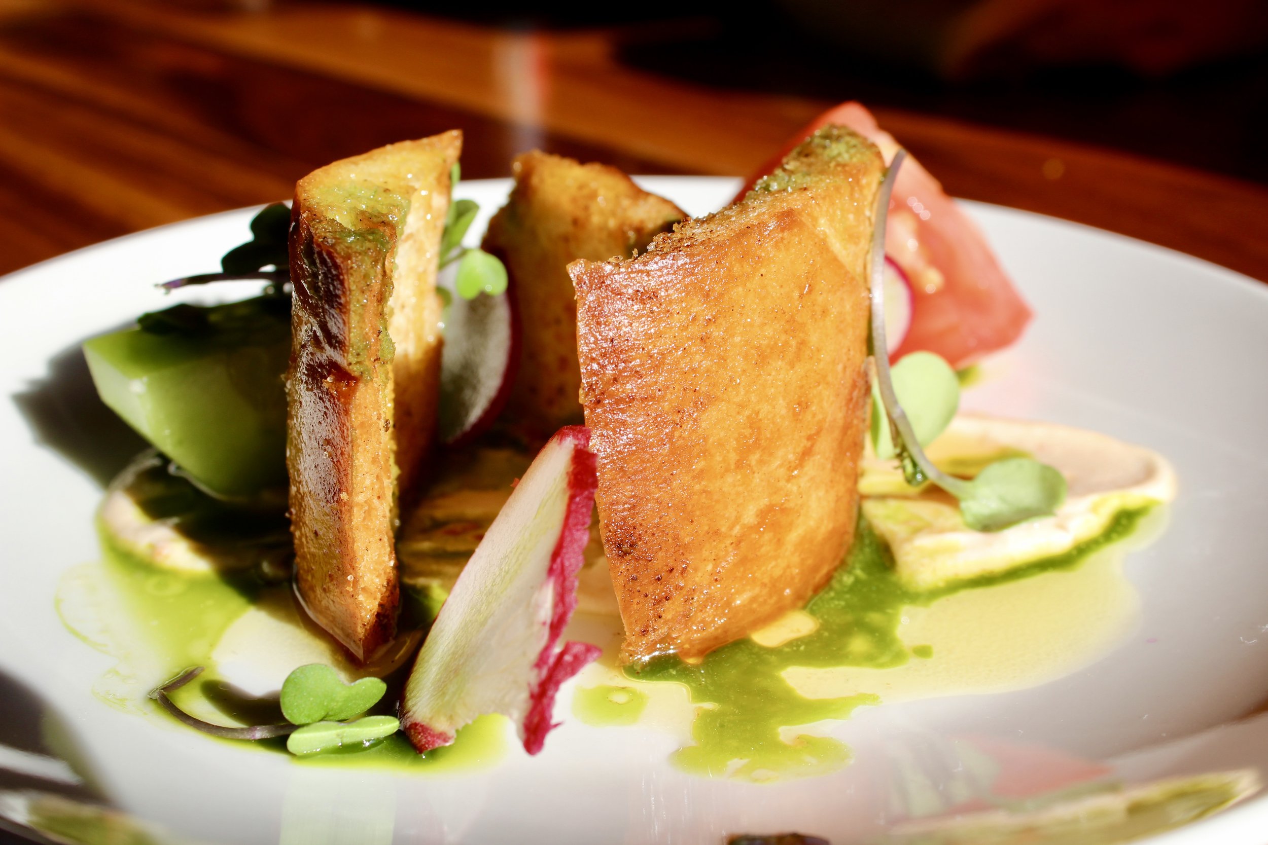 Close-up of an artfully plated gourmet dish featuring crispy fried potato wedges, sliced vegetables, microgreens, and a vibrant green sauce on a white plate.