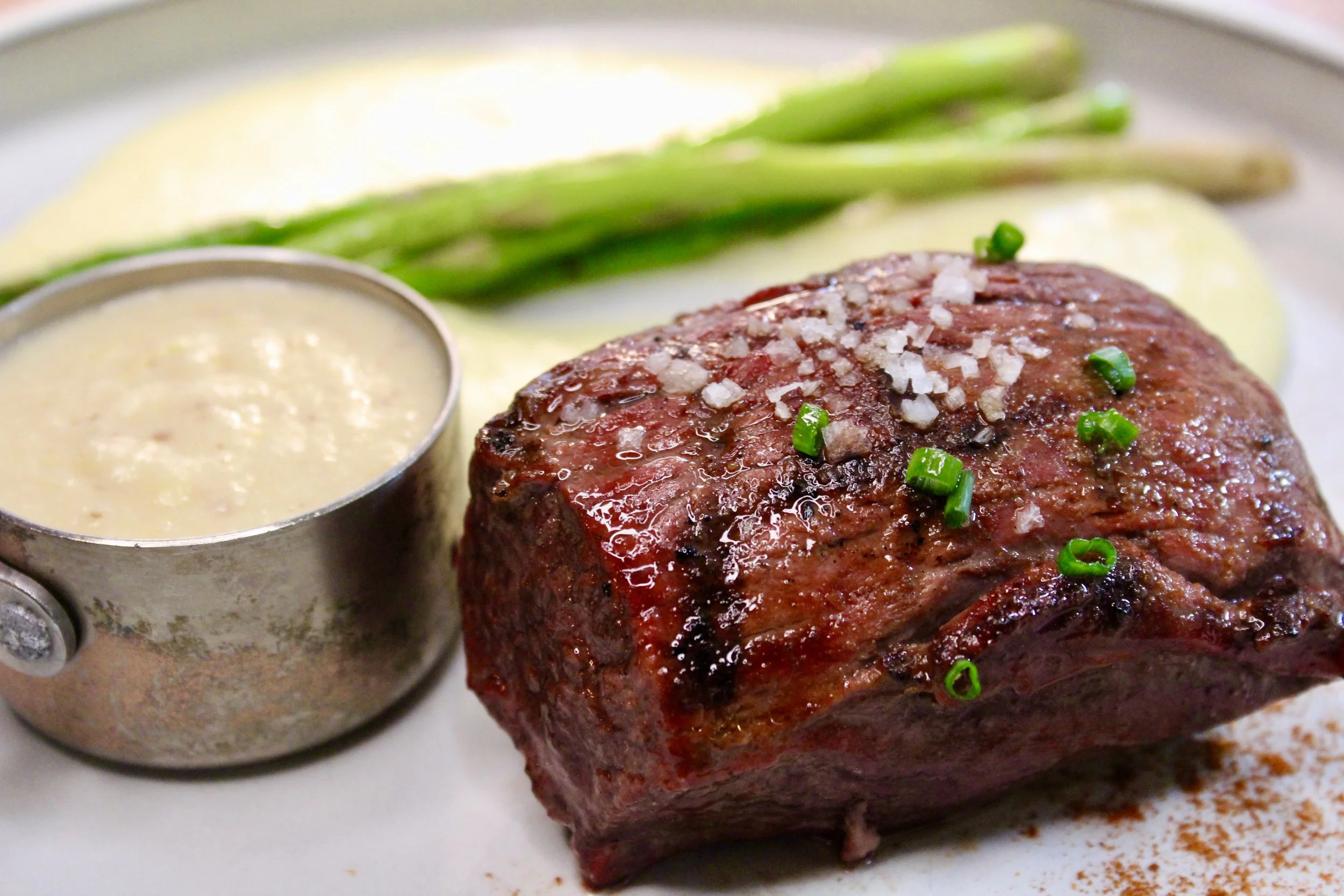 A plate with a cooked filet mignon steak topped with chopped onions and green onions, served with asparagus, mashed potatoes, and a side of creamy sauce.