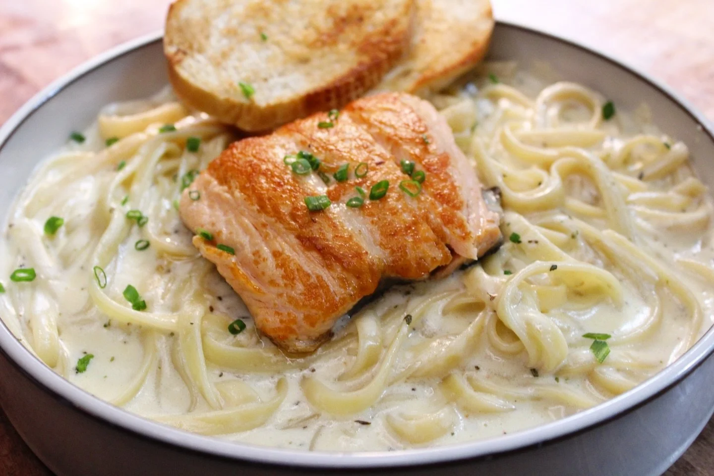 Creamy fettuccine alfredo with a grilled chicken breast topped with chopped green onions, served with garlic bread on a plate.