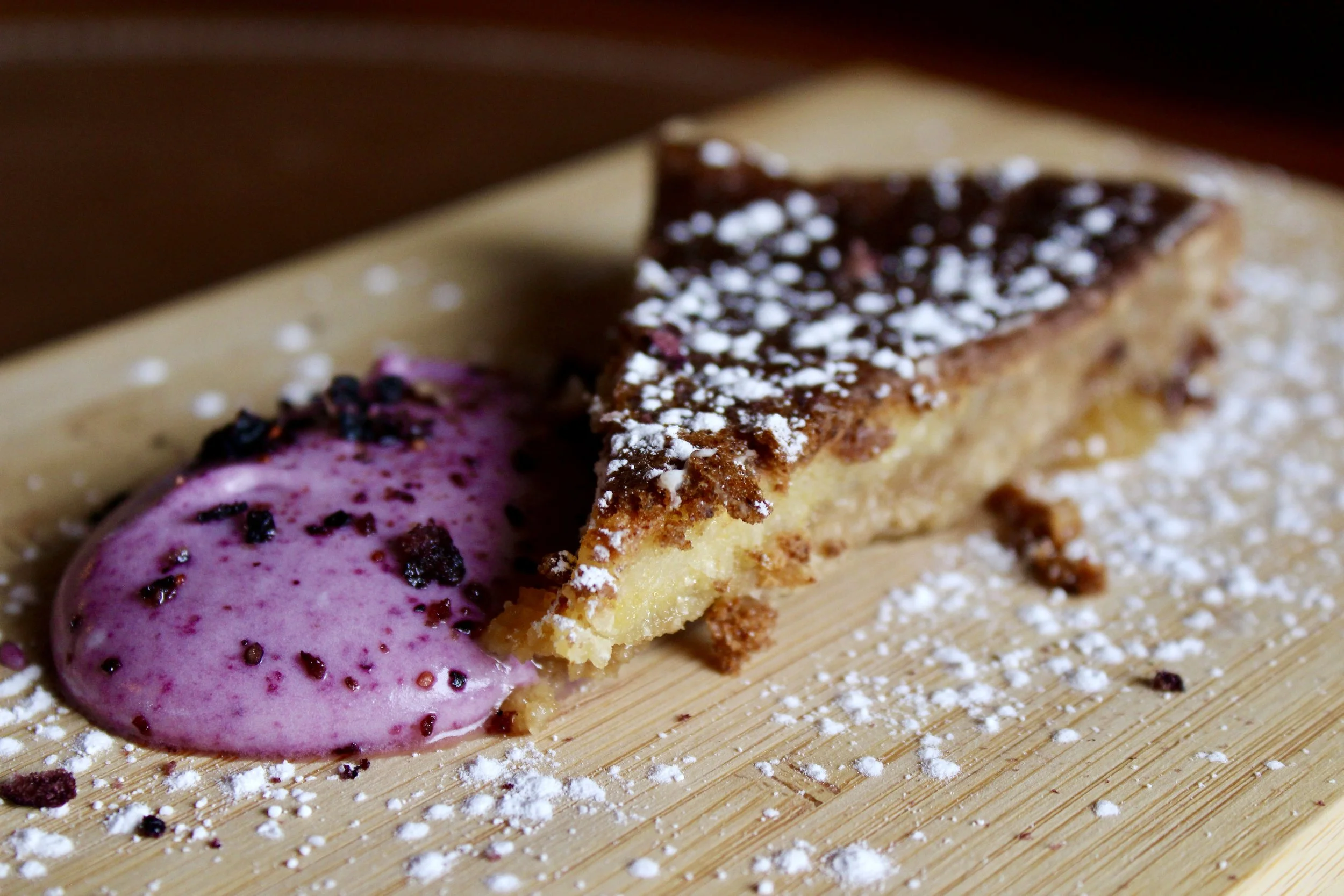 Slice of peanut butter pie with purple berry sauce and powdered sugar on a wooden surface.