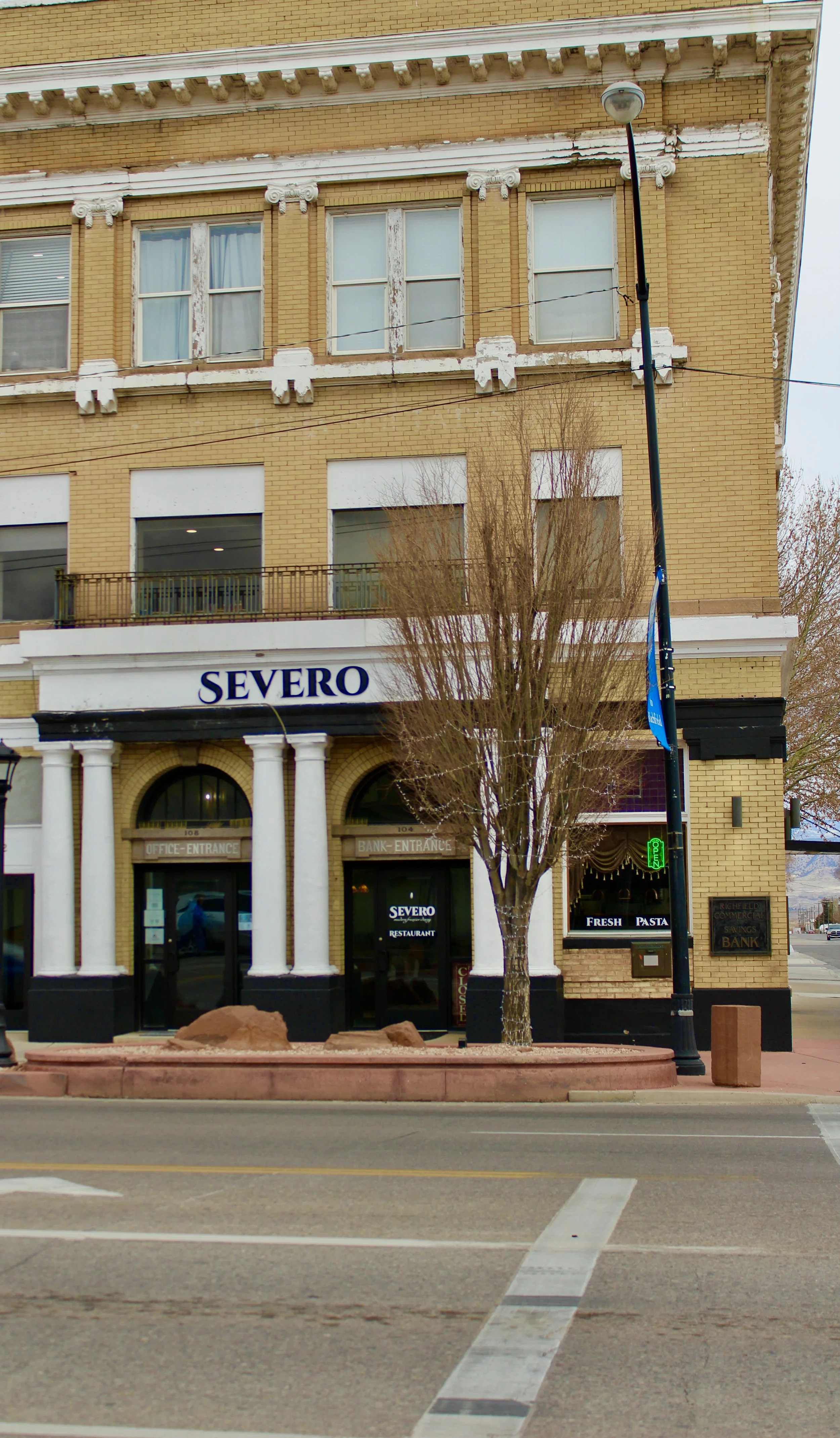 A four-story brick building with white decorative architectural elements, housing the Severo restaurant on the ground floor. The restaurant has white columns at the entrance, a sign with blue letters, and a tree in front. The street shows marked lanes and a pedestrian crosswalk.