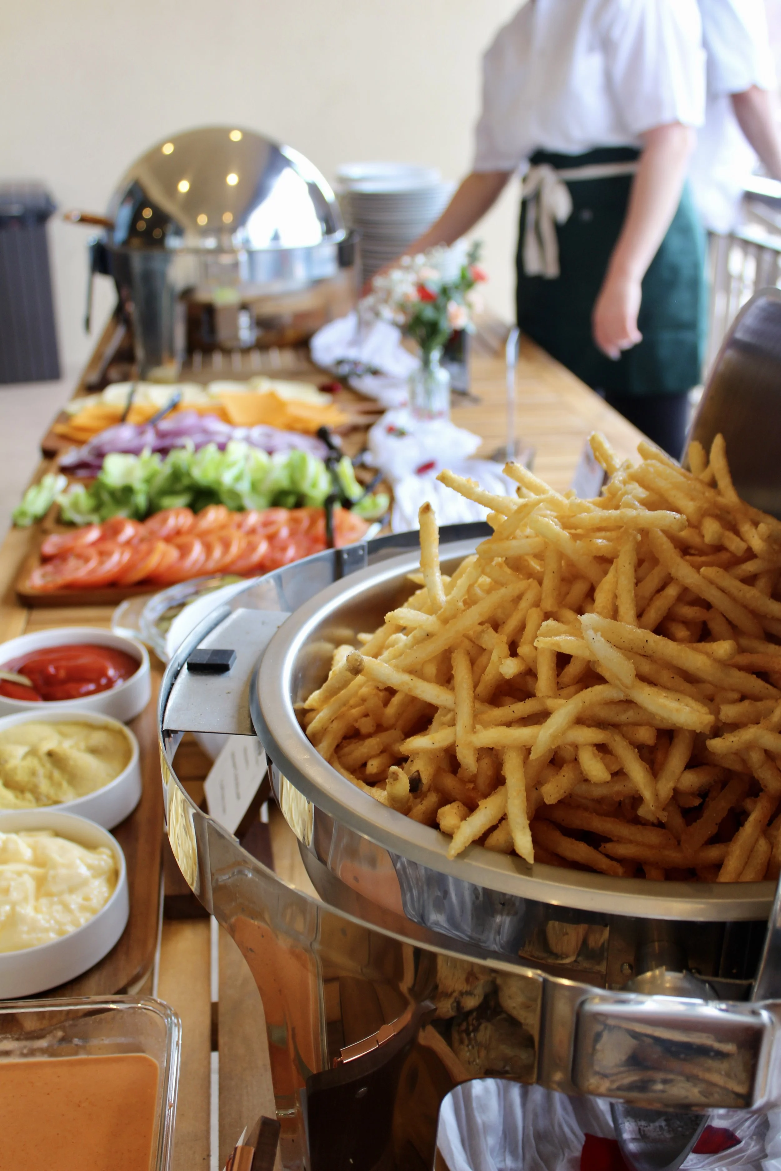 Fresh French fries in a large metal container on a buffet table with condiments and sliced vegetables in the background.