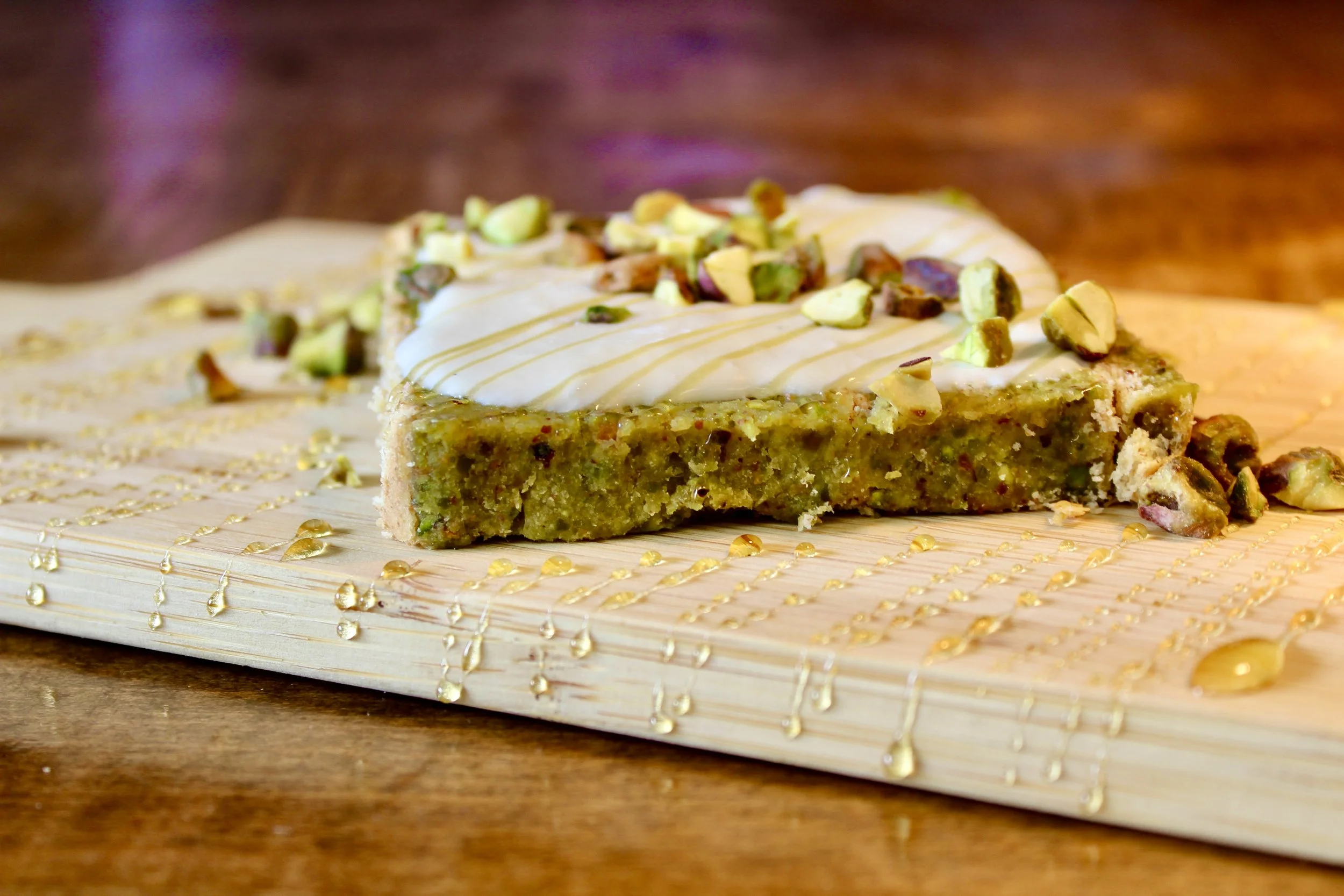 Slice of pistachio cake with white icing, topped with chopped pistachios, on a wooden board with gold drizzle.