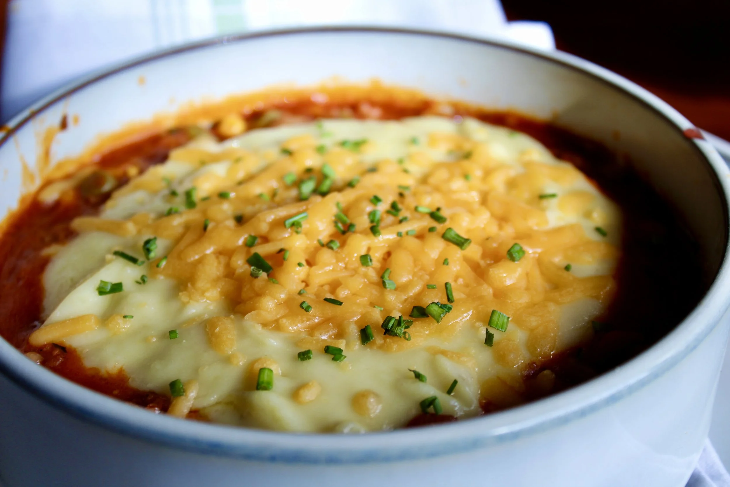 Baked cheesy casserole with melted cheese, chopped chives, and tomato sauce in a white bowl.