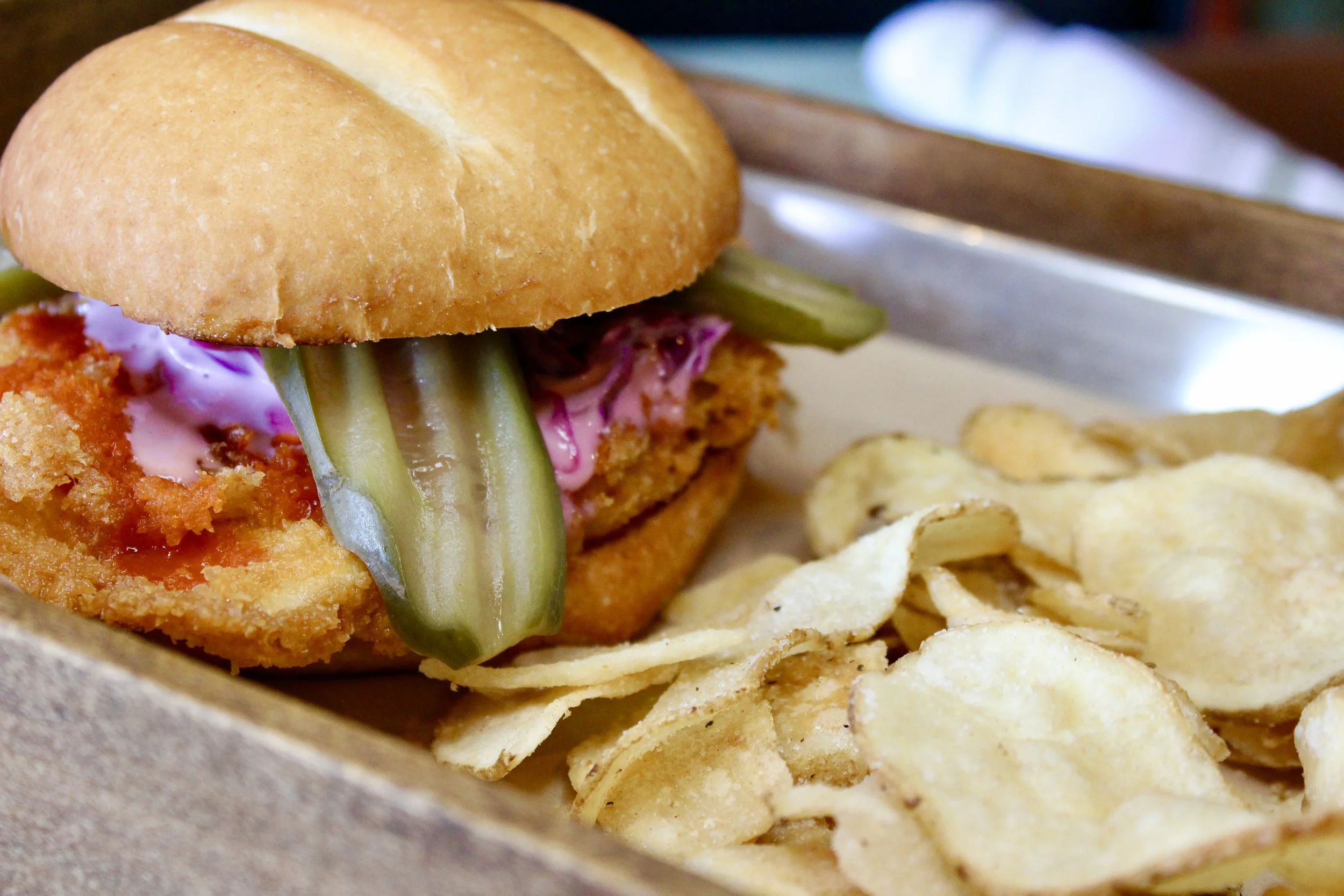 A close-up of a fried fish sandwich with pickles and pink sauce, served with potato chips.
