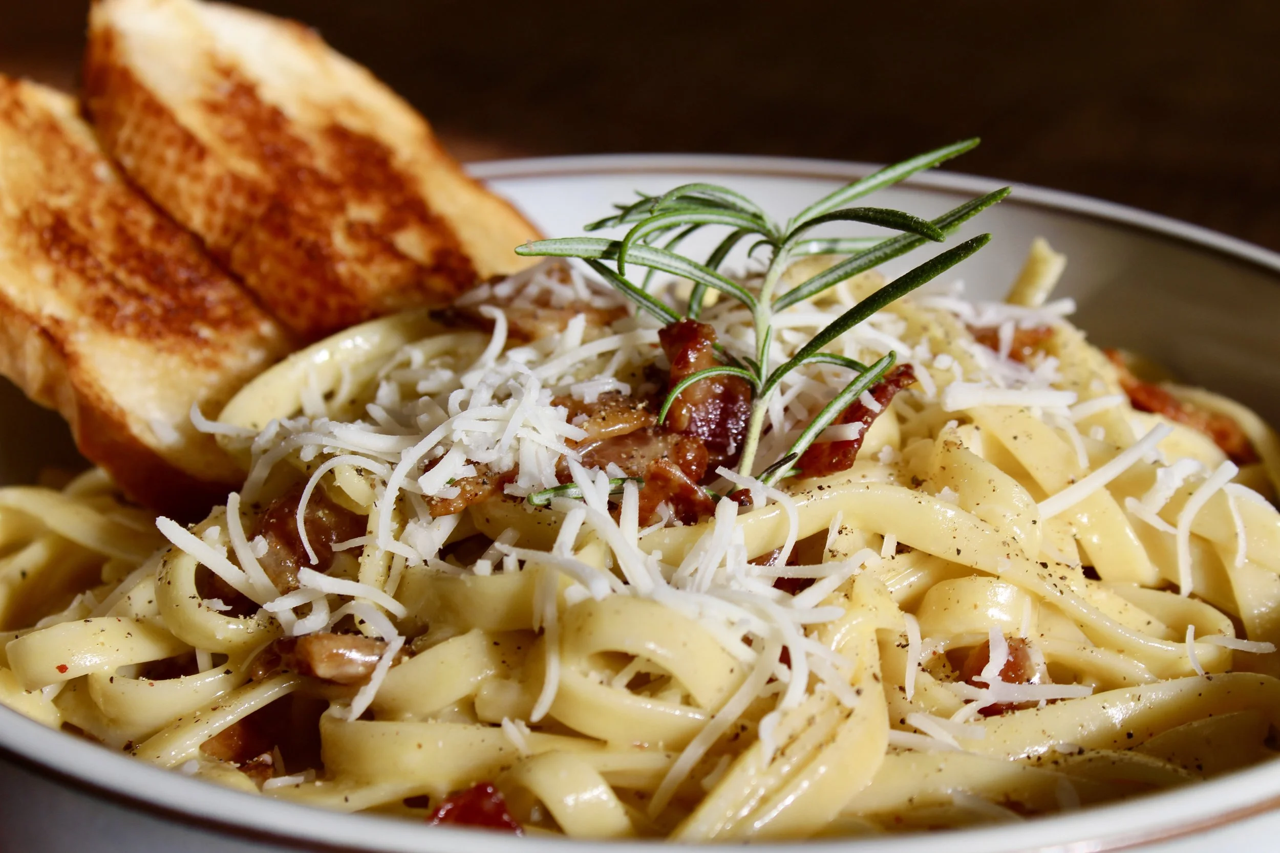 Close-up of a bowl of cheesy pasta topped with shredded cheese, bacon bits, and a sprig of rosemary, with toasted bread slices on the side.