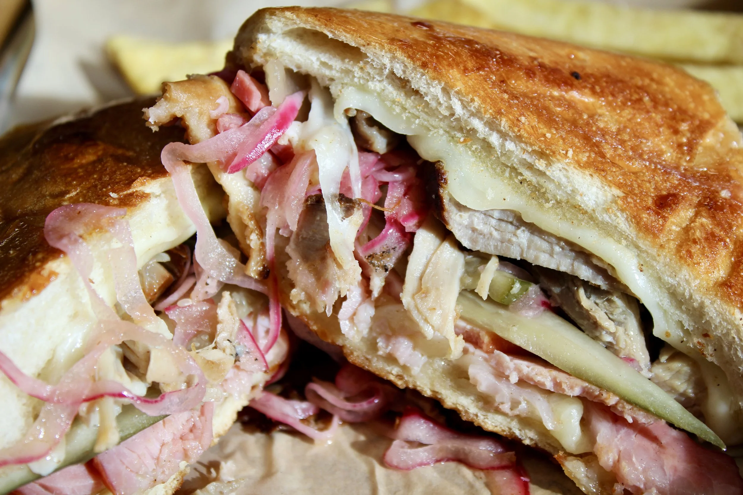 Close-up of a sandwich with crispy toasted bread, sliced turkey, melted cheese, pickles, and coleslaw, with French fries in the background.