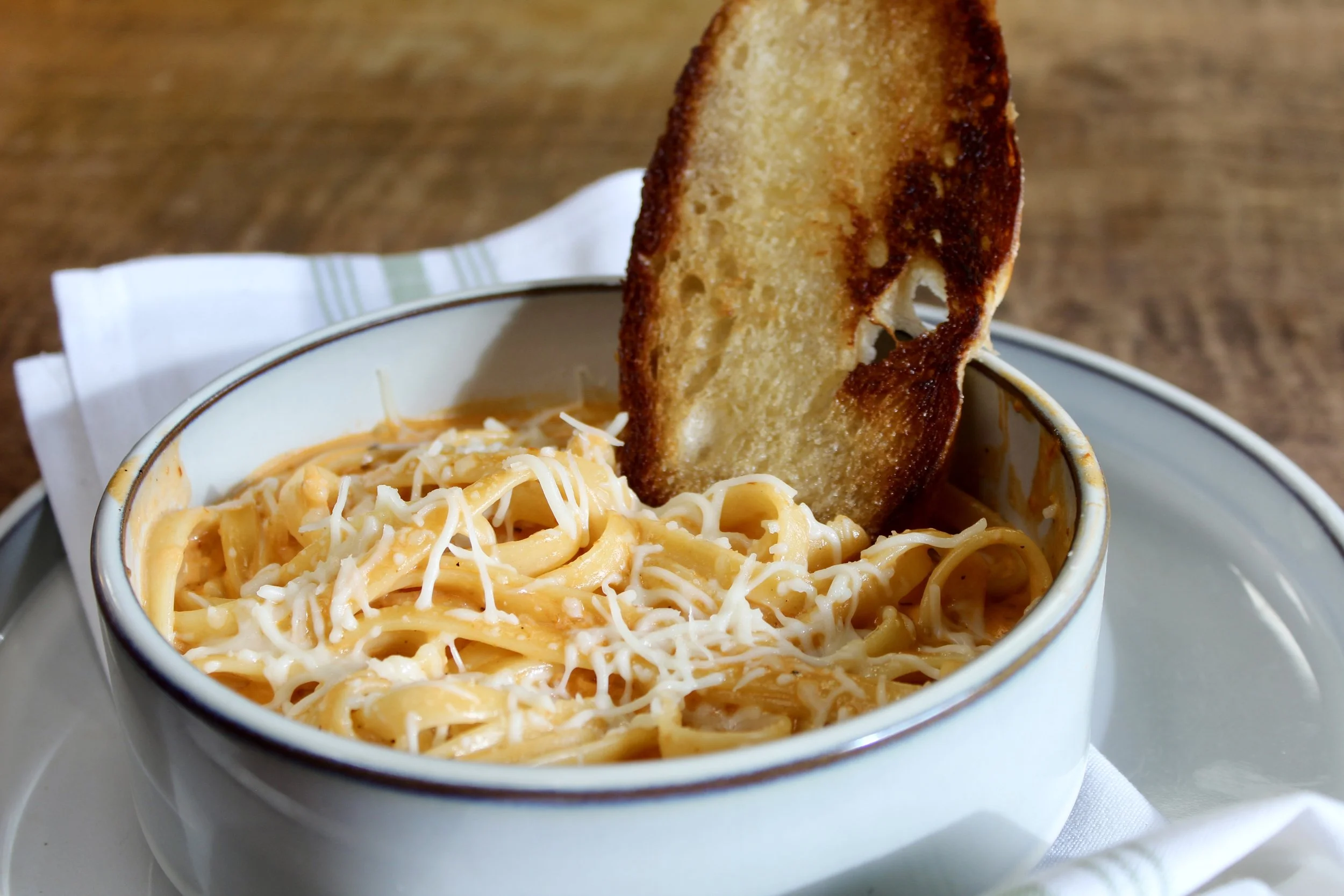 A bowl of creamy Alfredo pasta topped with shredded cheese, served with a slice of toasted bread on a white plate.