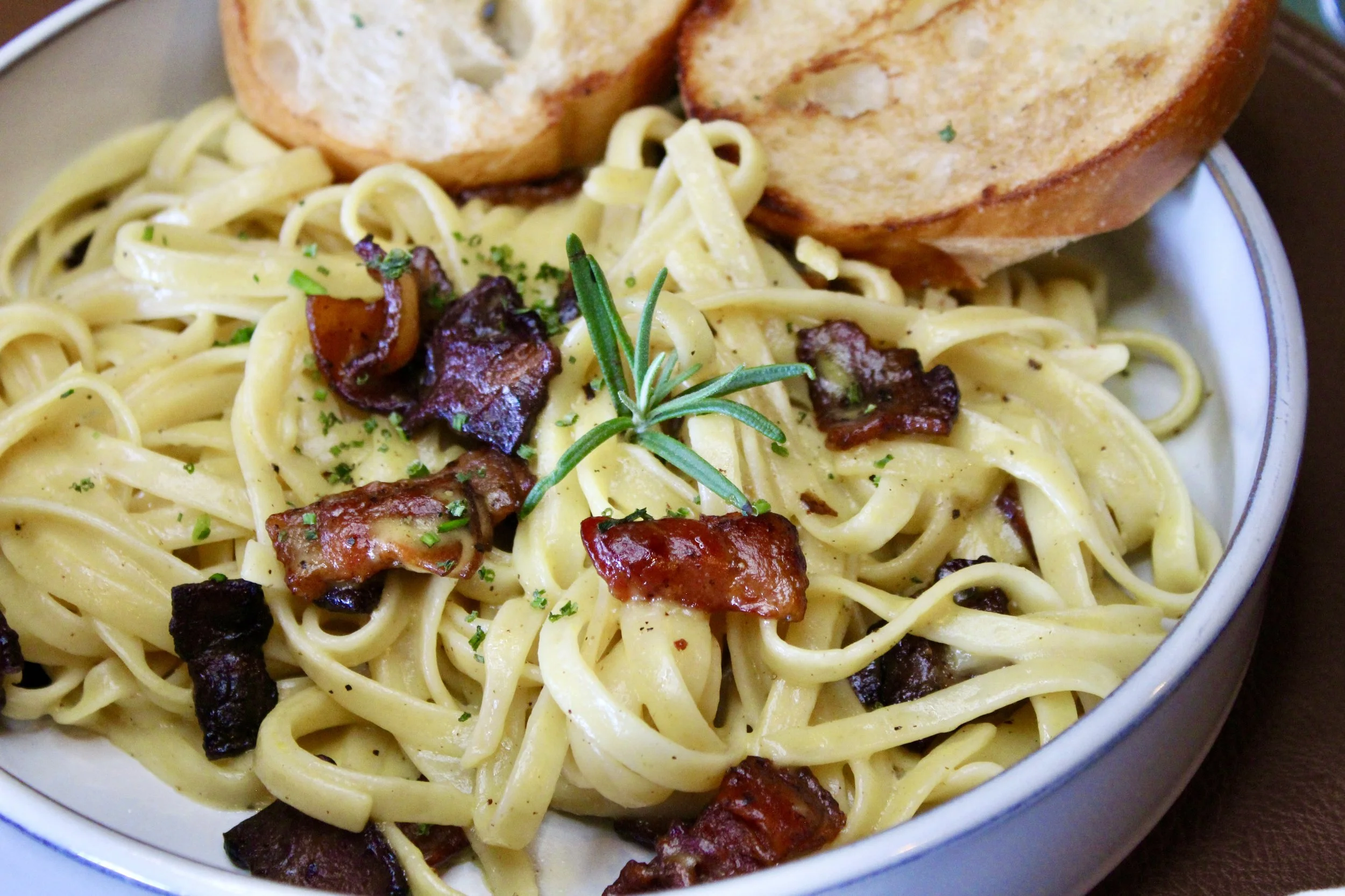 Close-up of a bowl of creamy fettuccine pasta with bacon bits, garnished with a sprig of rosemary, served with toasted bread slices.