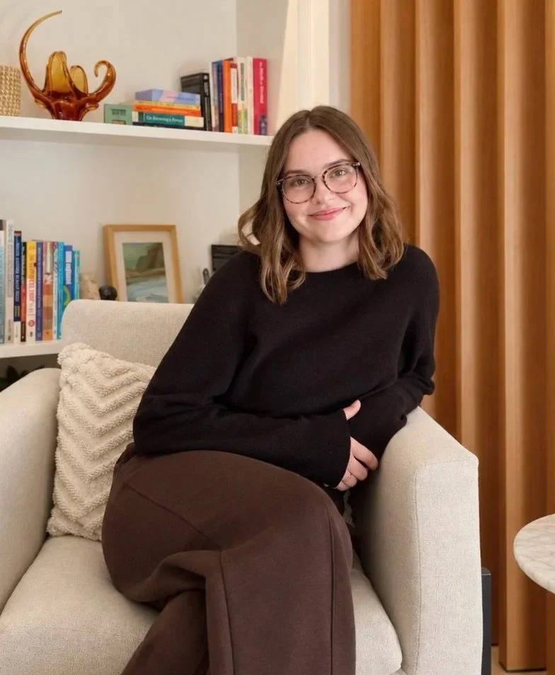 A young woman with shoulder-length brown hair, wearing glasses and a black sweater, sitting on a beige sofa in a cozy living room with bookshelves in the background.