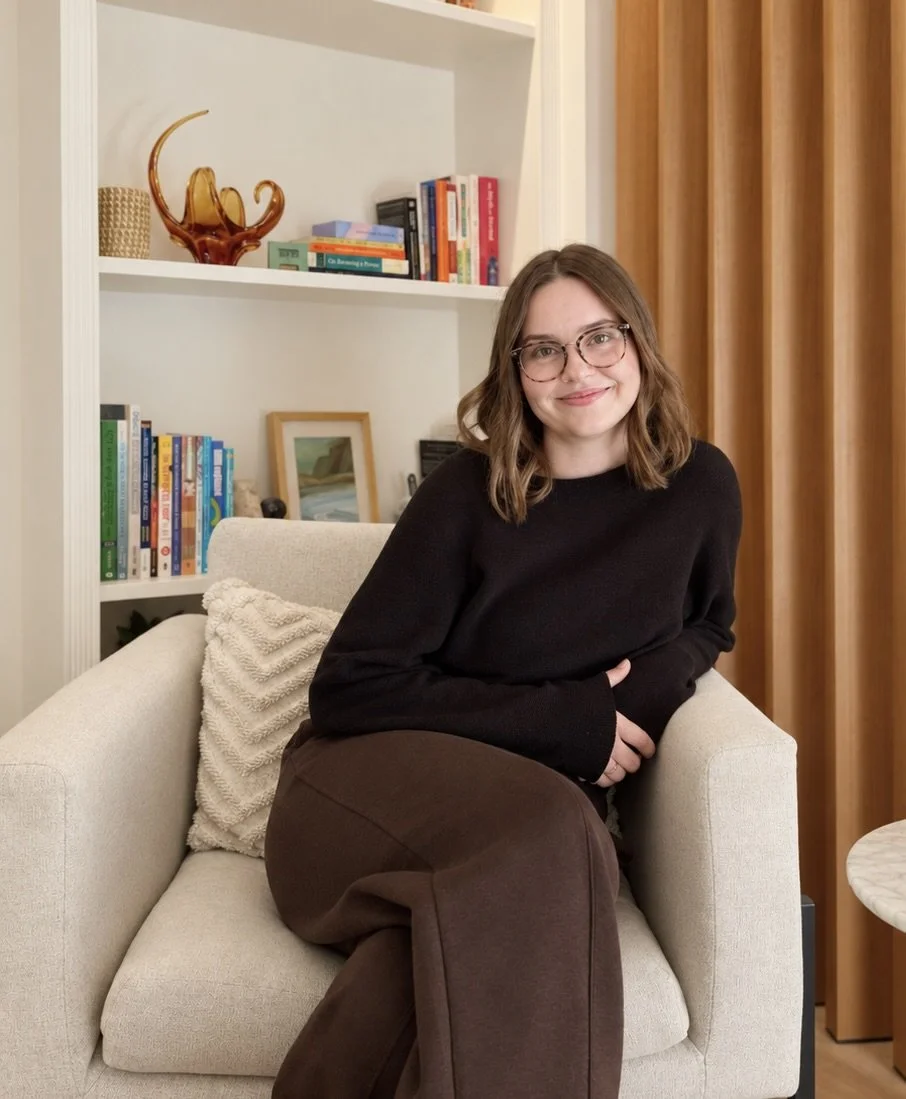 A woman with glasses, shoulder-length brown hair, wearing a black sweater, sitting on a beige armchair in a cozy room with a bookshelf behind her.