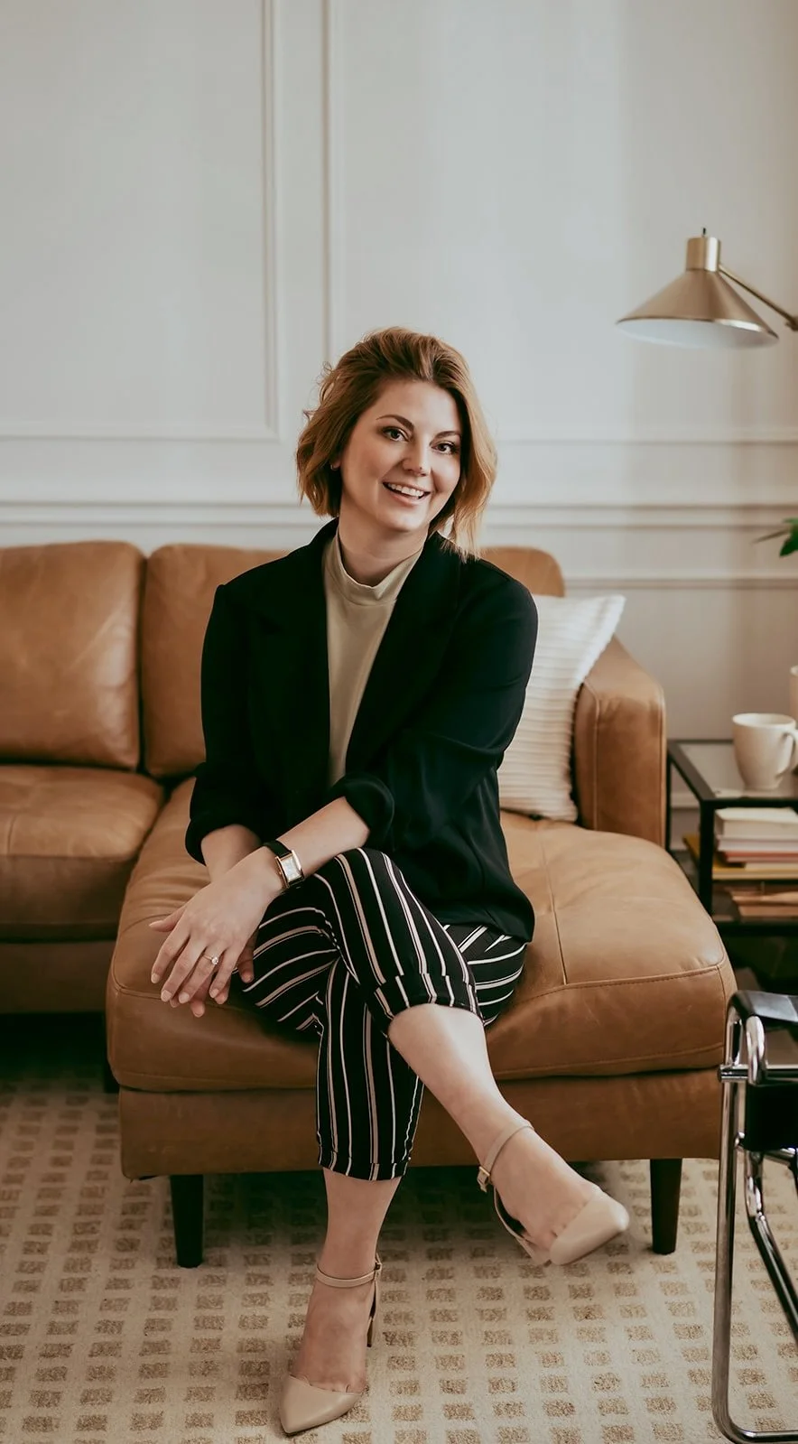 A smiling woman with shoulder-length reddish-brown hair sitting on a tan leather couch in a cozy modern living room. She is a psychologist who offers in-person and virtual therapy for adults. Her office is in Edmonton, AB.