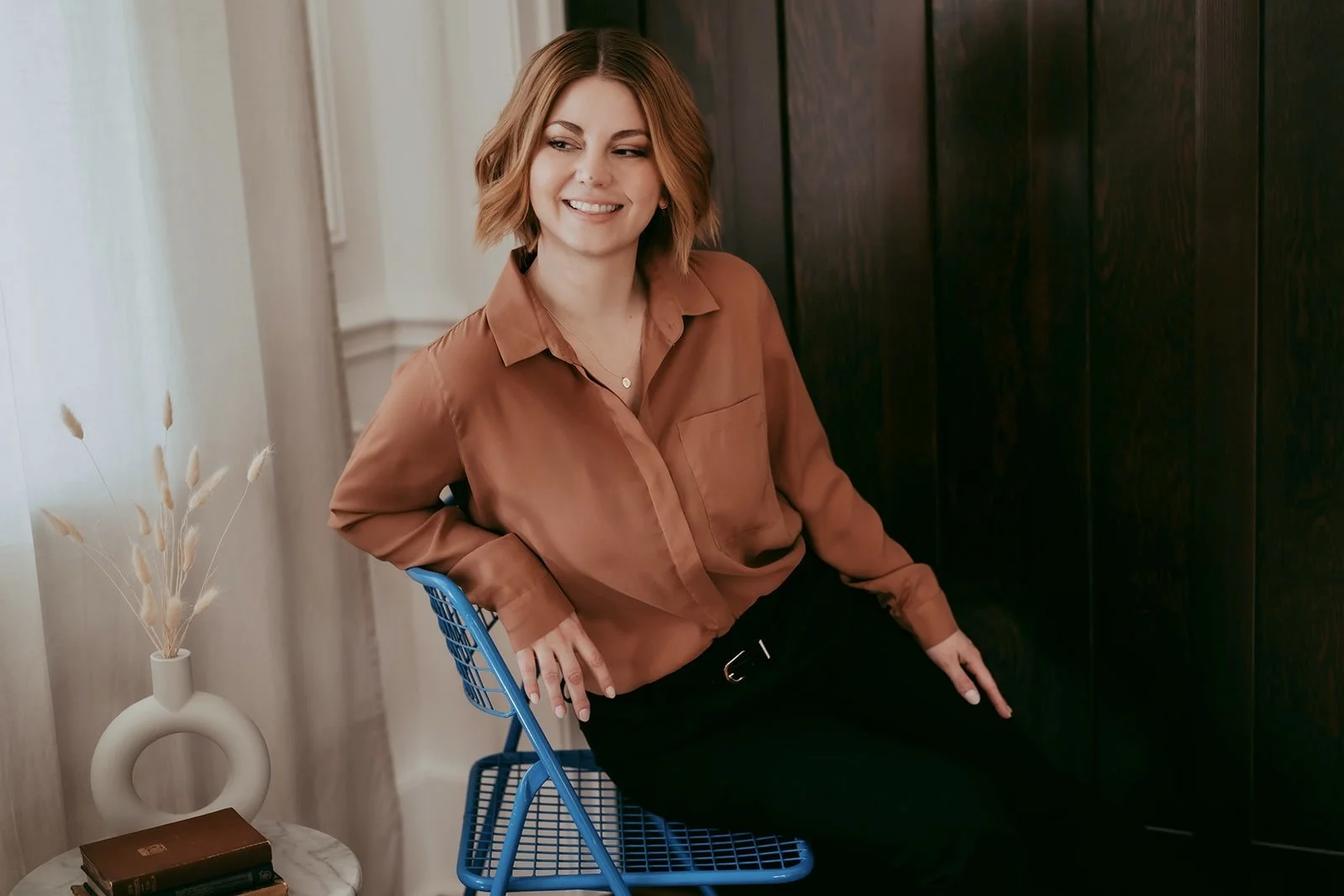 A woman with shoulder-length brown hair sitting on a blue wire chair, smiling, wearing a brown blouse and black pants, indoors near a dark wooden wall, with a white vase containing dried grasses on a small table beside her.
