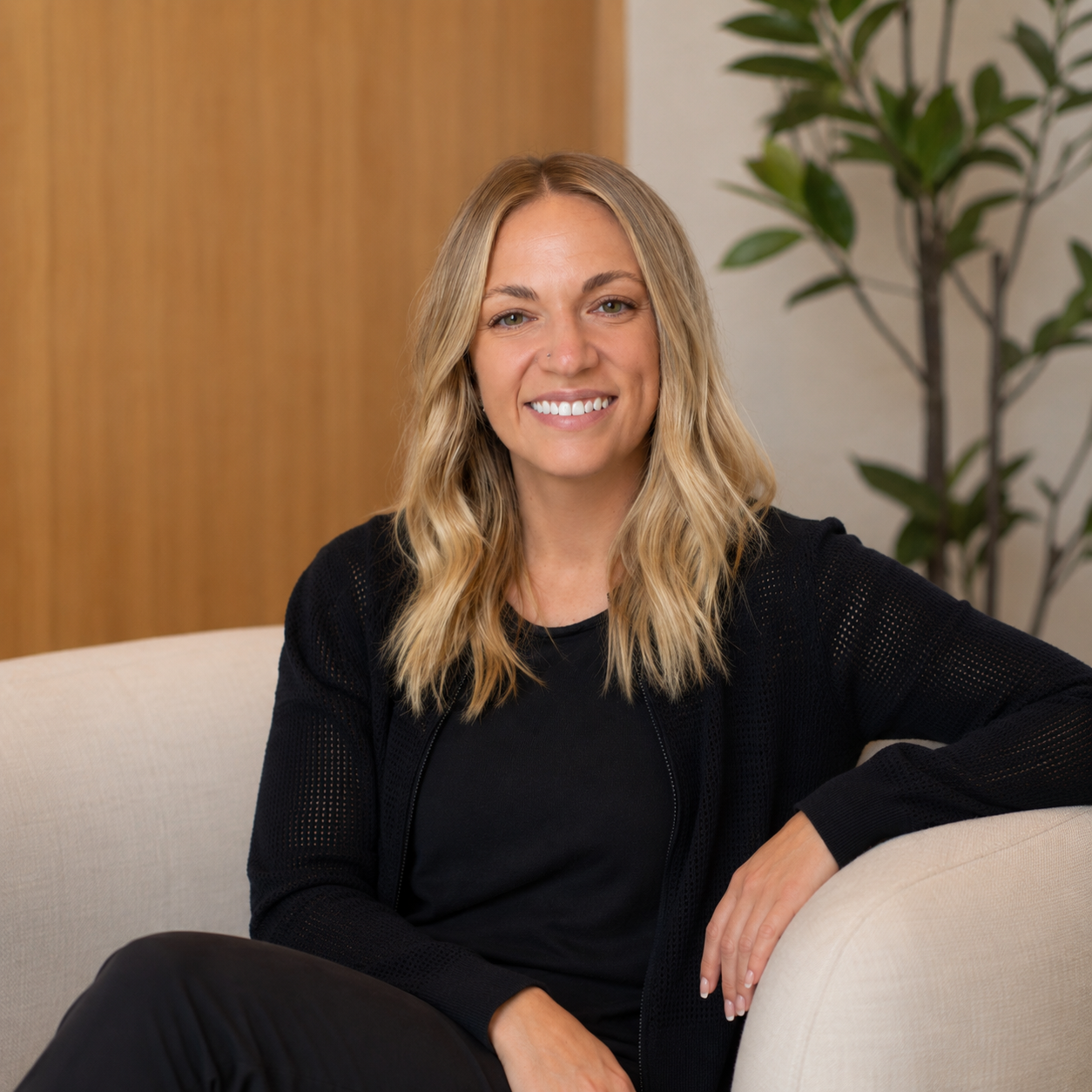 A woman with shoulder-length blonde hair smiling, sitting on a beige couch, wearing a black top and a black cardigan, with a houseplant and wooden wall in the background.