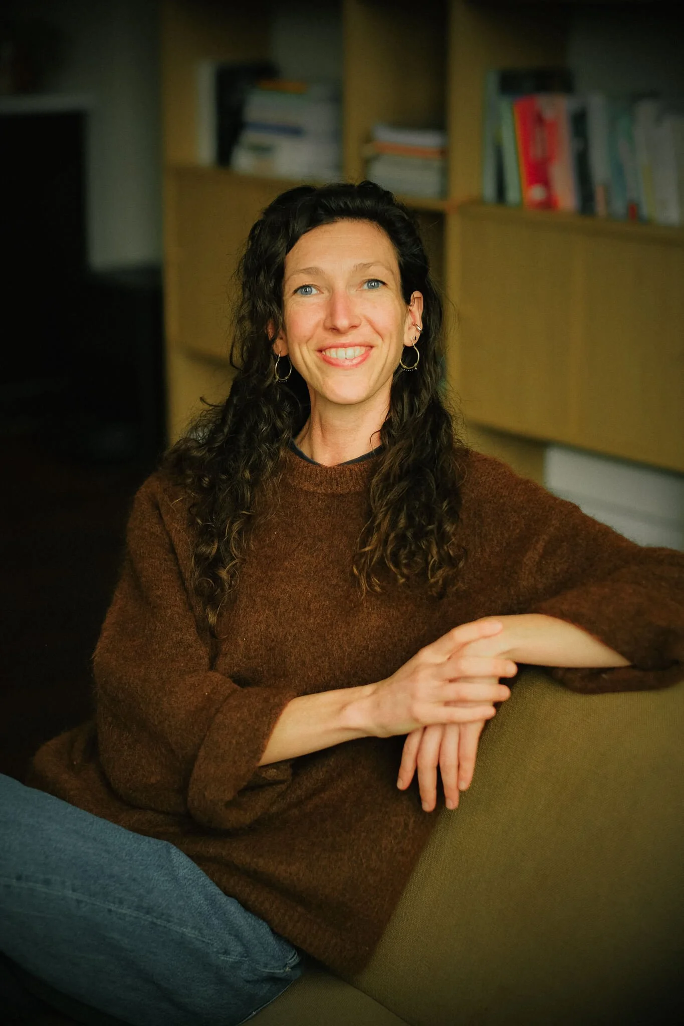 Een vrouw met lang, krullend haar en blauwe ogen, lachend en zittend op een bank in een kamer met boekenkasten op de achtergrond.