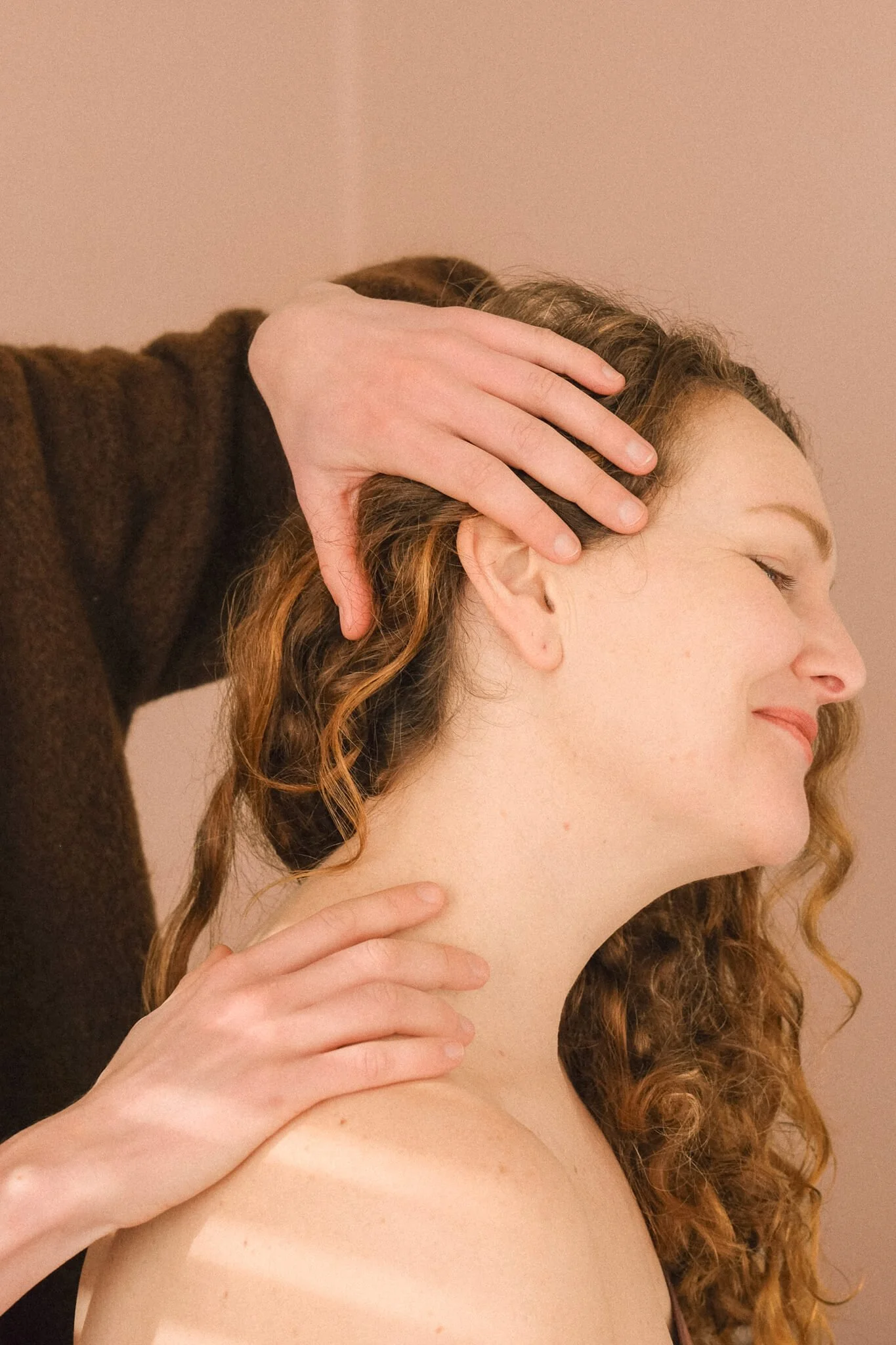 Close-up van een vrouw met lange, krullende, roodbruin haar die wordt gemasseerd, met een zachte, roze achtergrond.