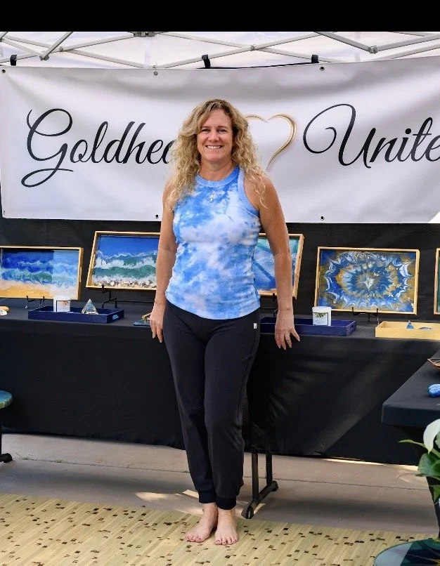 A woman with curly blonde hair smiling, standing barefoot in front of an art display at an outdoor event, with a banner in the background reading 'Goldheart United'.