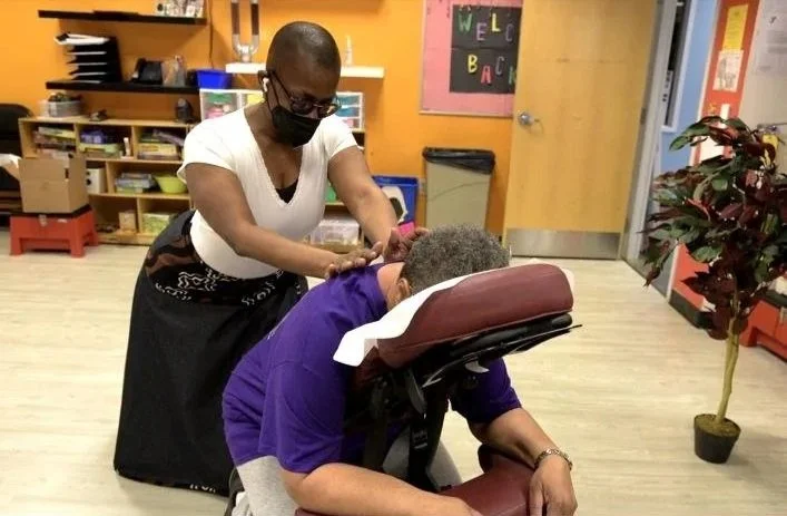 A person receiving a massage from a massage therapist in a room with shelves, a plant, and colorful walls.
