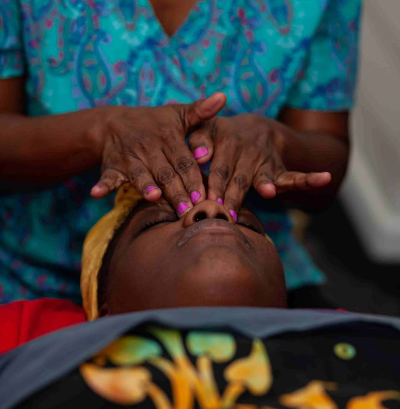 A person receives a facial massage from a therapist with painted pink nails, wearing a colorful shirt and a yellow headband.
