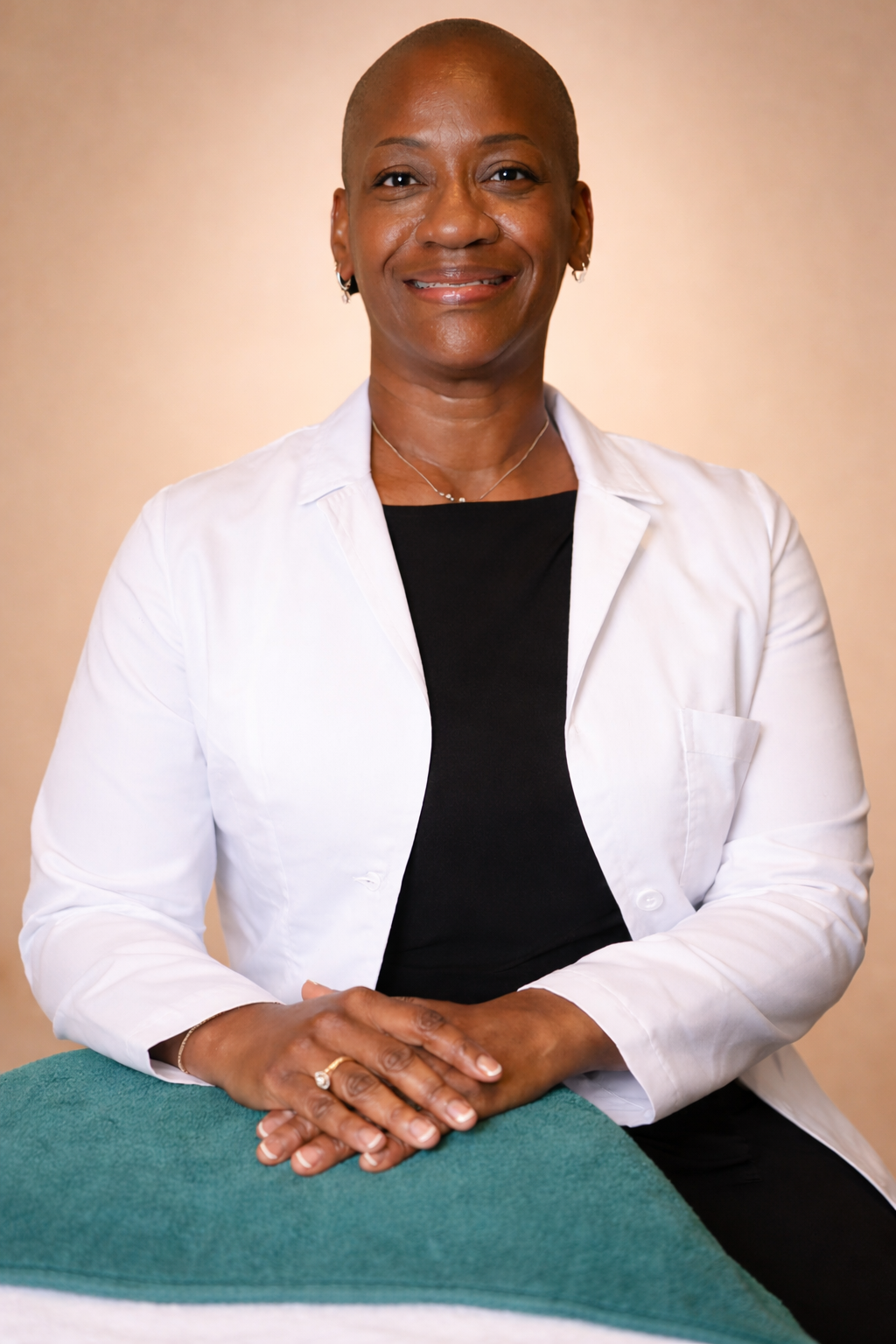 A professional woman with a shaved head, wearing a white lab coat over a black top, sitting at a table with folded hands, smiling, against a plain beige background.
