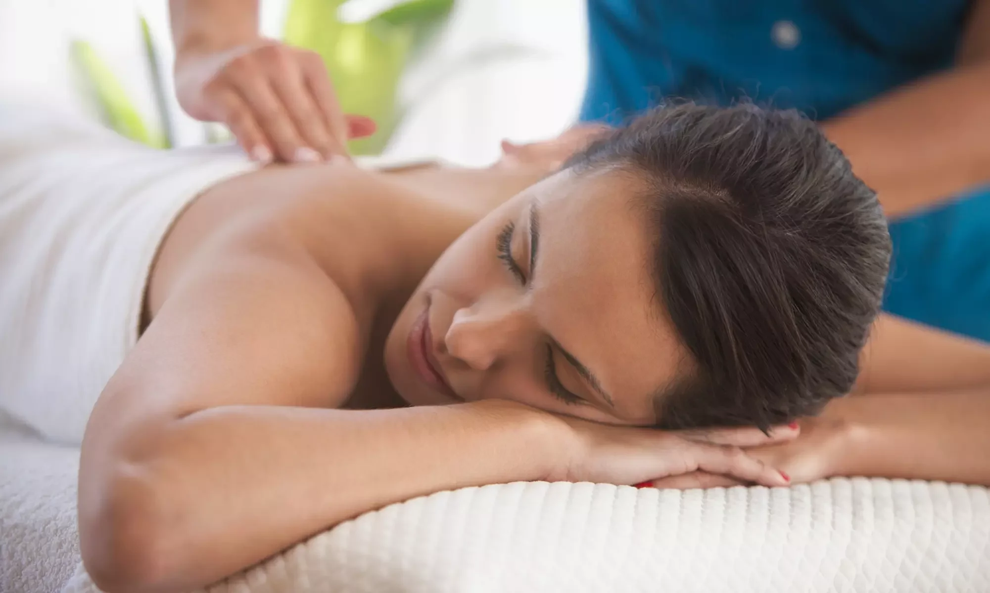A woman receiving a massage while lying face down with eyes closed, resting her head on her hands, in a bright, relaxing setting.