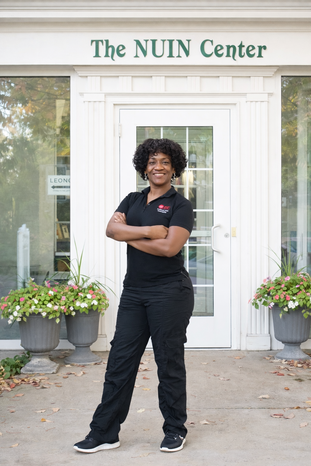A woman with curly black hair stands confidently with her arms crossed in front of a building labeled 'The NUIN Center.' She is wearing a black polo shirt and black pants.
