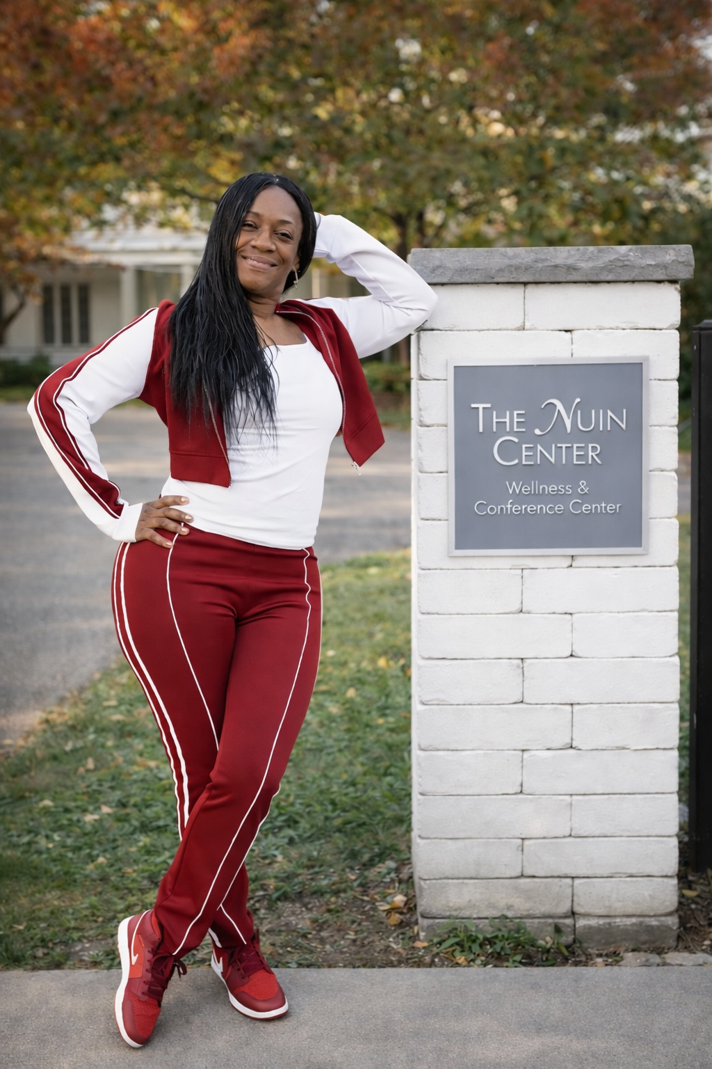 A woman in red athletic pants, a white shirt, and a red jacket stands with one hand on her hip and the other resting on her head next to a sign for The Nunn Center Wellness & Conference Center. She is outdoors with autumn trees in the background.