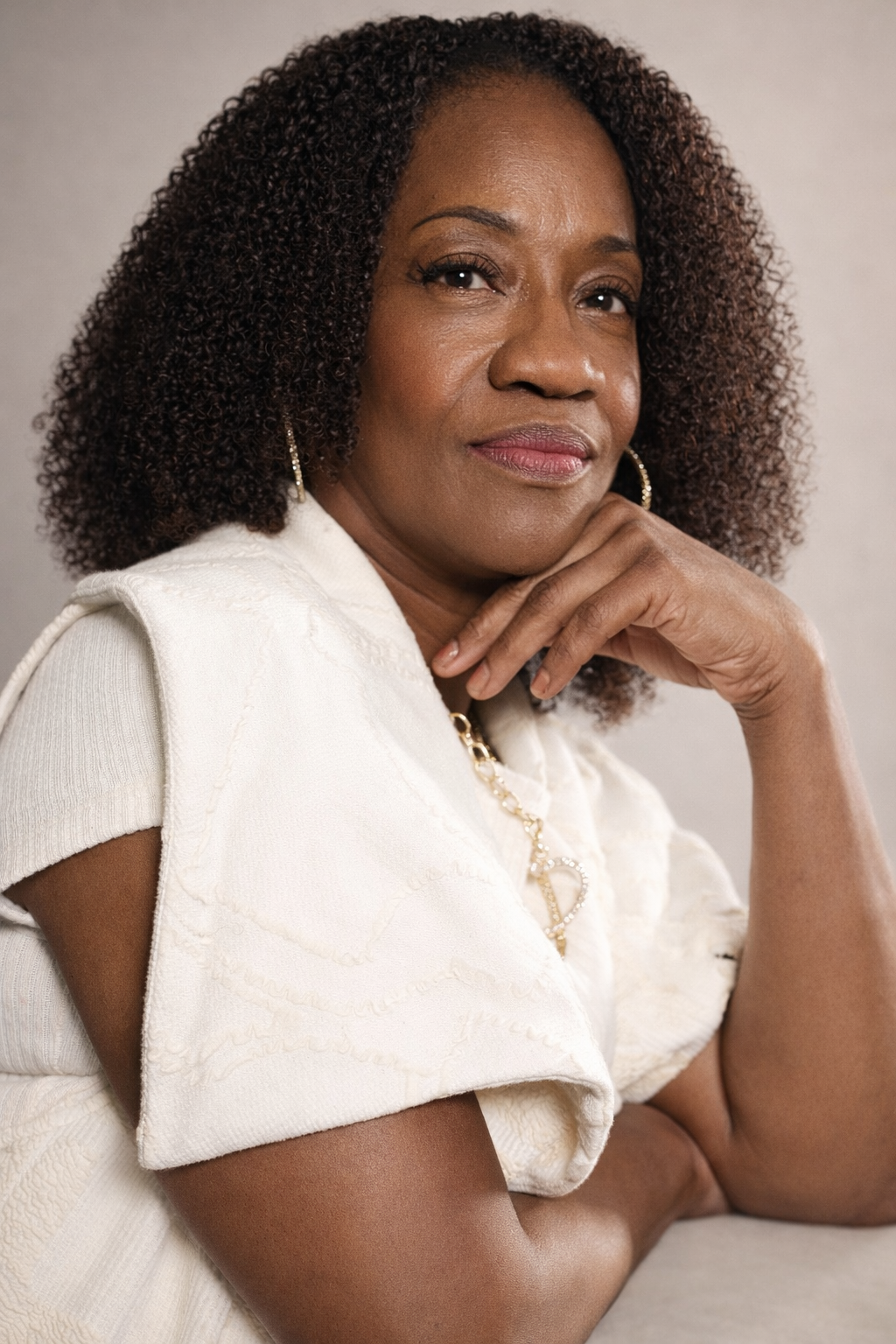 A confident African American woman with curly hair, dressed in white, resting her chin on her hand, looking directly at the camera.