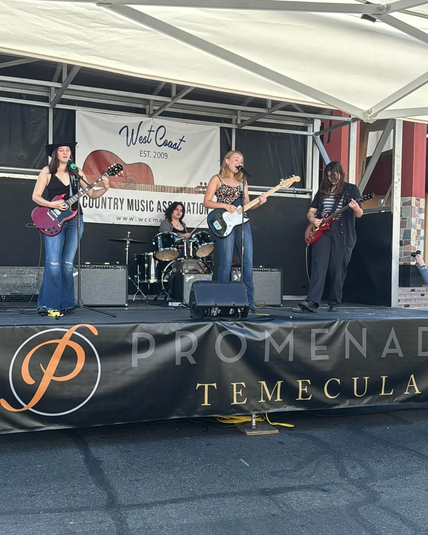 A four-member band performing on an outdoor stage under a white canopy, with a banner behind them reading 'West Coast  Country Music Association'. The band includes a lead singer with long blonde hair playing bass, a guitarist with dark hair and a wide-brim hat, another guitarist with long dark hair, and a drummer with curly dark hair. The stage has a black banner with 'PROMENADE TEMECULA' written on it.