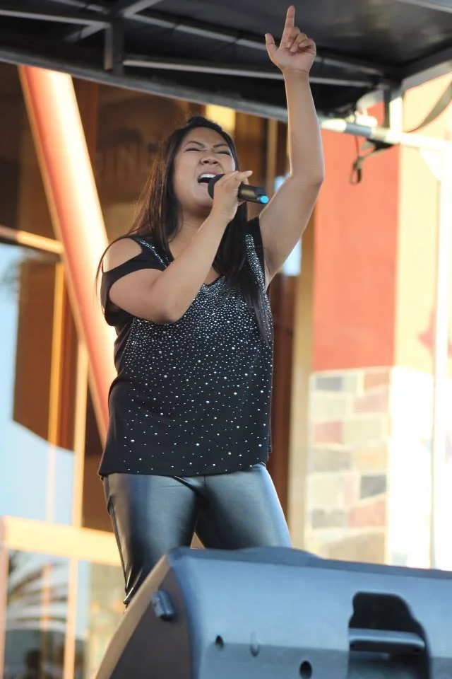 A female singer performs passionately on the WCCMA Hot August stage, holding a microphone in her right hand, eyes closed, and her left hand raised with her index finger pointing upward. She is wearing a black, sparkly top and shiny black pants.