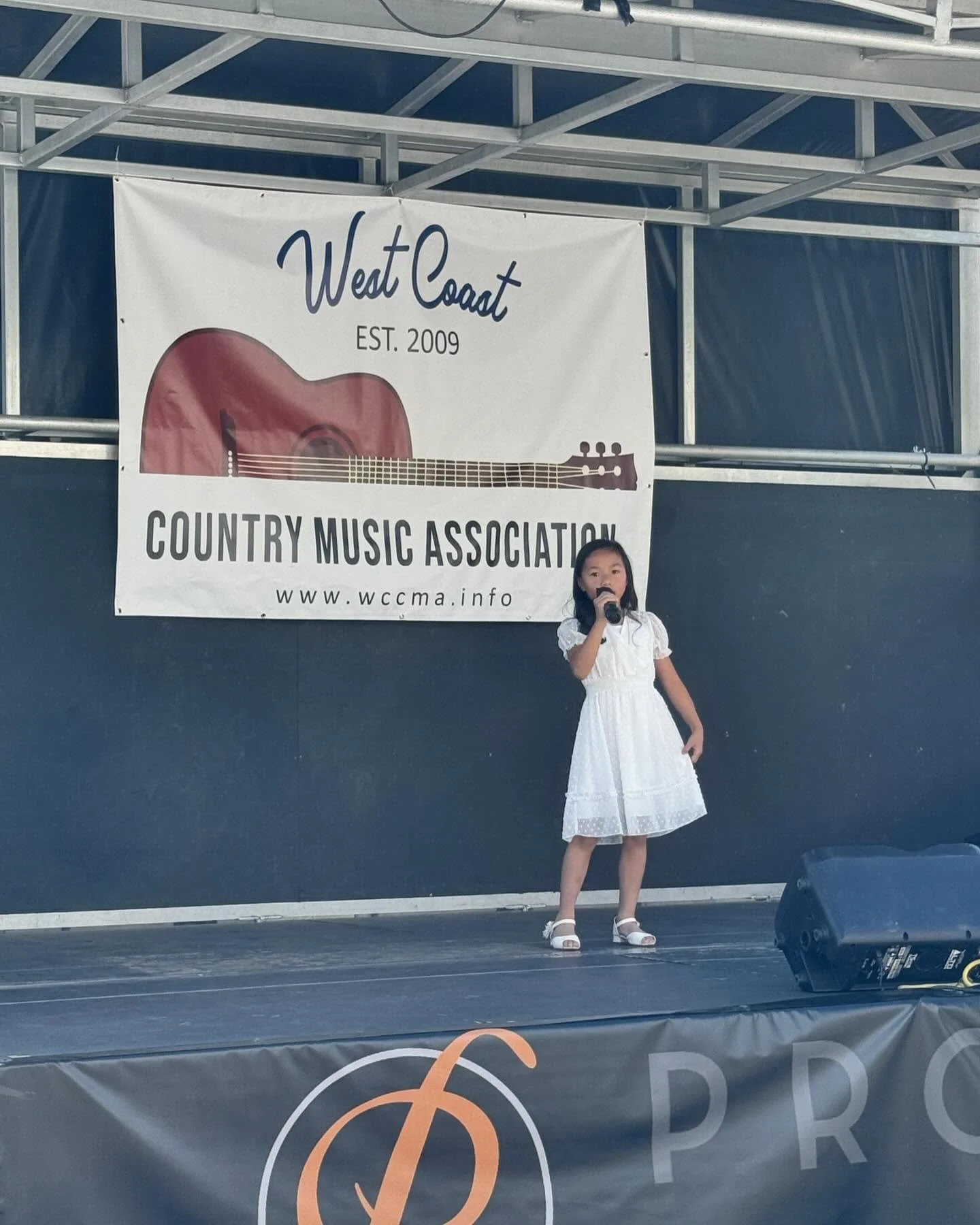 A young girl in a white dress holding a microphone on stage at the annual Hot August country music competition, with a large banner behind her that reads 'West Coast Country Music Association EST. 2009", and features a guitar illustration.