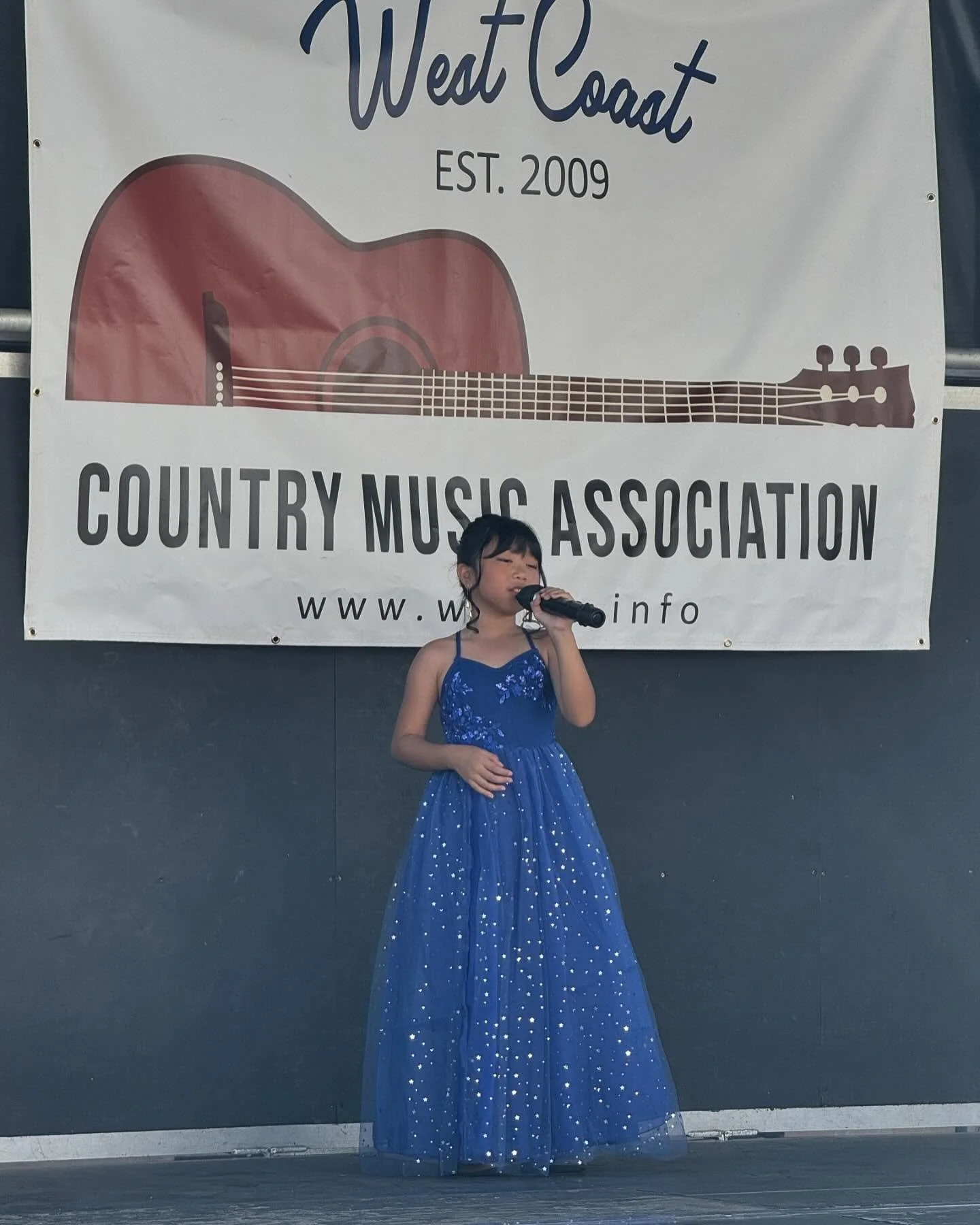 A young girl in a blue dress holding a microphone on stage at the annual Hot August country music competition, with a large banner behind her that reads 'West Coast Country Music Association EST. 2009", and features a guitar illustration.