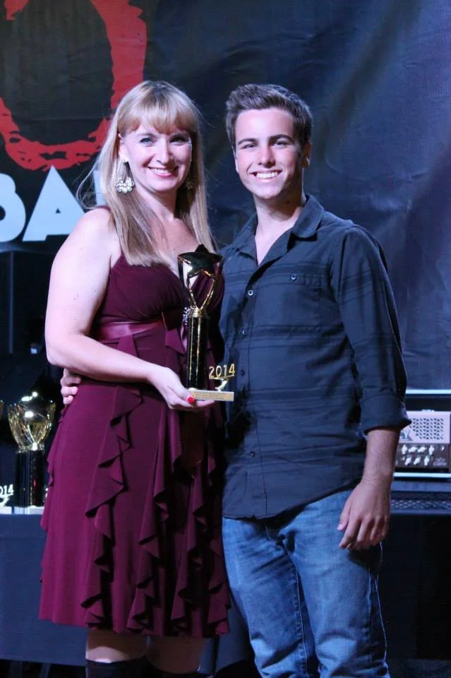 A woman and a young man standing together at the WCCMA Hot August Award Show, smiling, with awards and trophies in the background. The woman is holding a trophy and wearing a burgundy dress, and the young man is dressed in a black shirt and jeans.